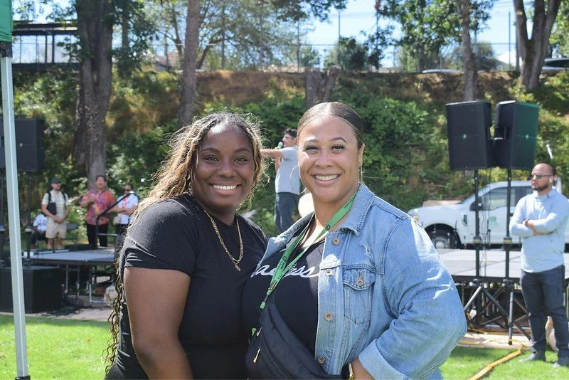 Two women smiling outdoors during a daytime event, with a stage and speakers in the background.