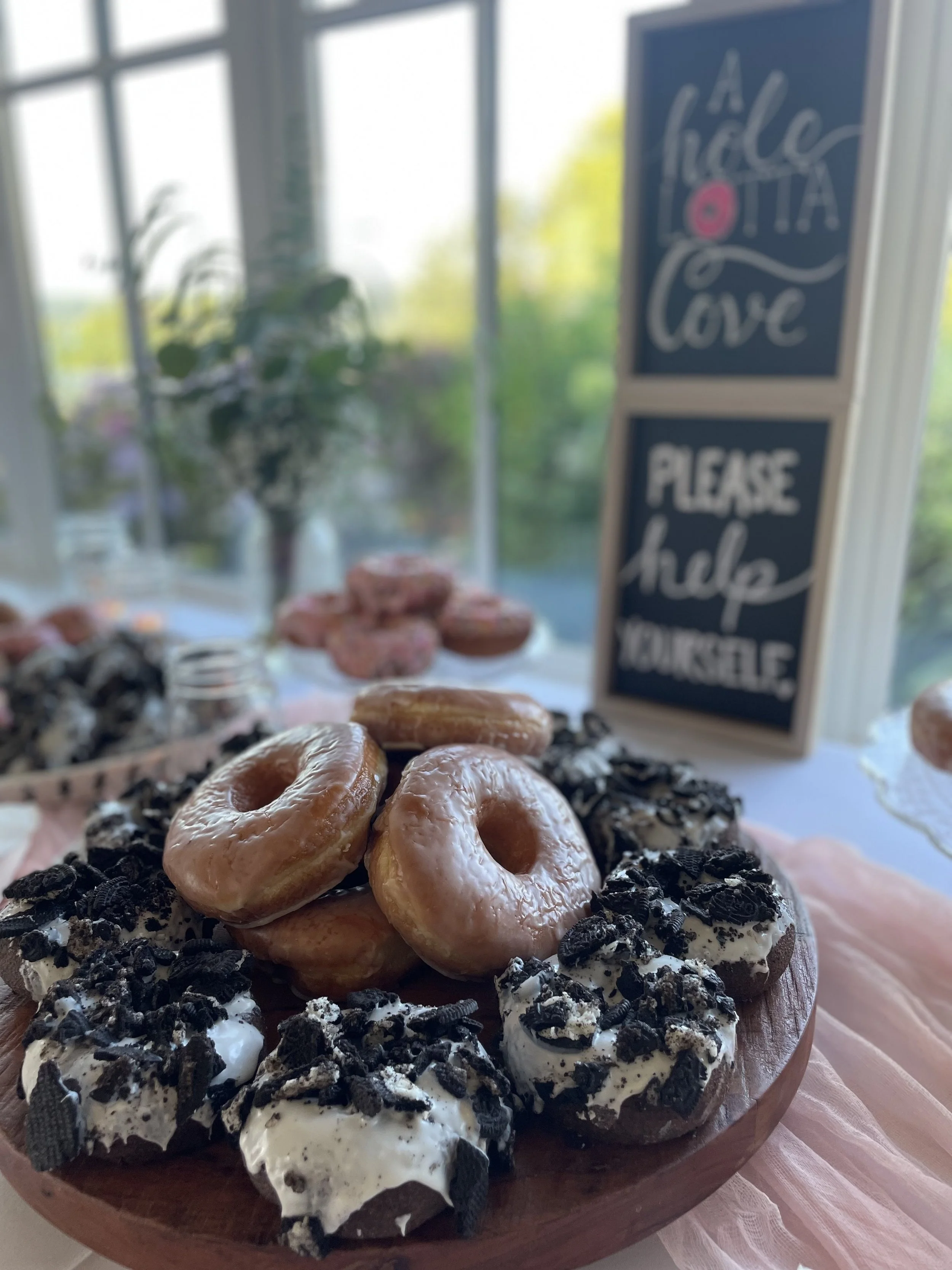 Assorted donuts on a wooden platter, with a blurred window and a chalkboard sign that reads 'A whole lotta love' and 'Please help yourself.'