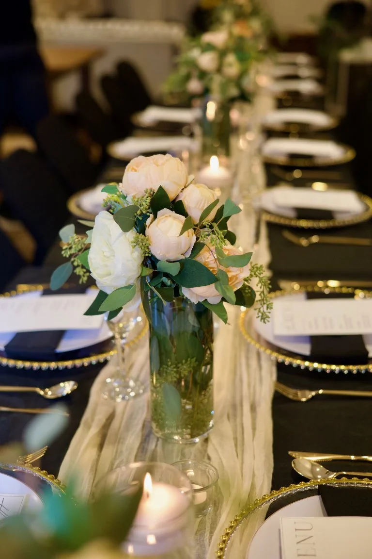 Elegant dining table setup with a tall floral centerpiece of white peonies and greenery, black and gold plates, white napkins, glassware, and a beige table runner in a dimly lit room.