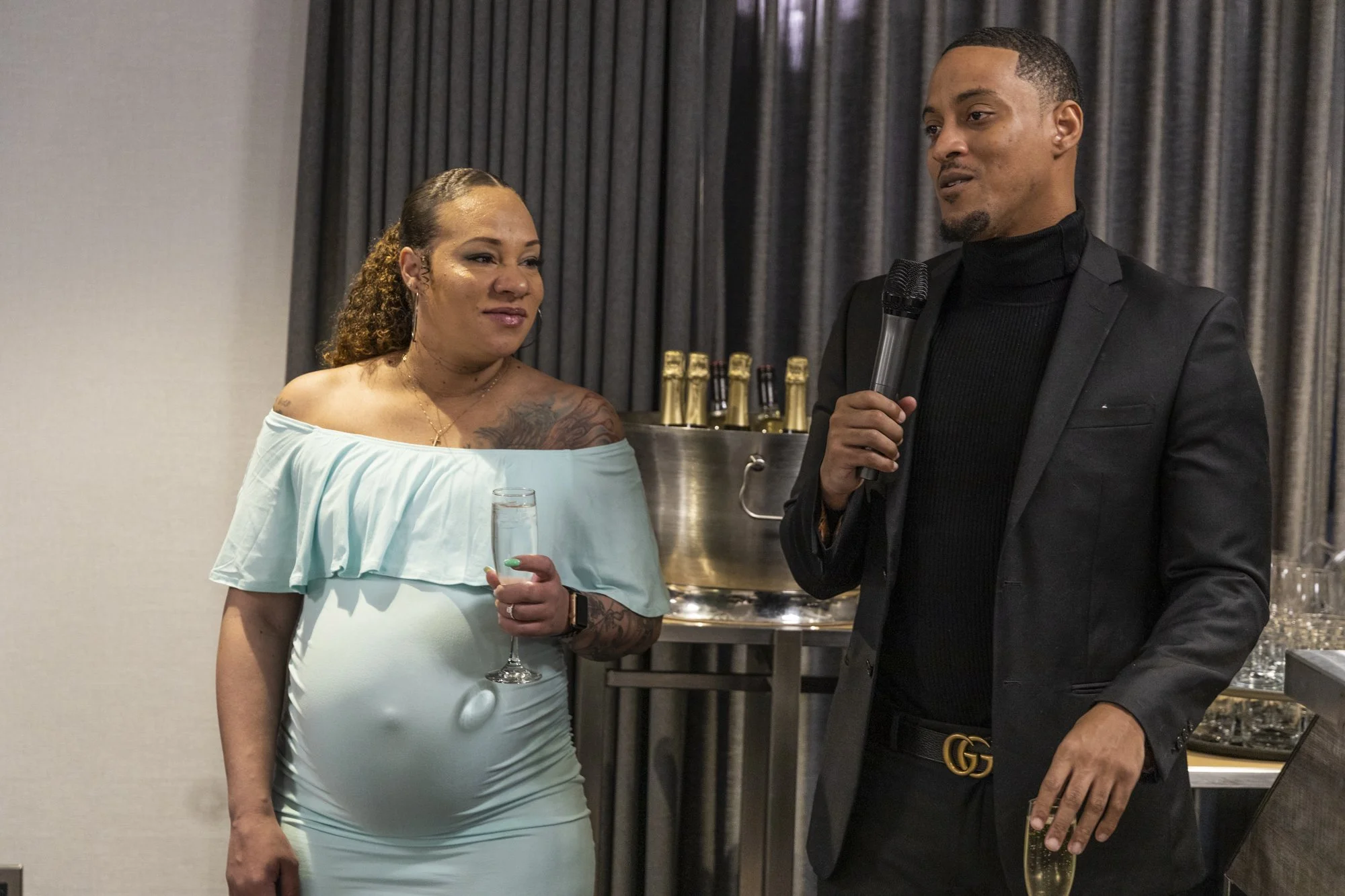 A woman in a light blue off-shoulder dress holding a glass of champagne looks at a man in a black suit holding a microphone at a social gathering or celebration.
