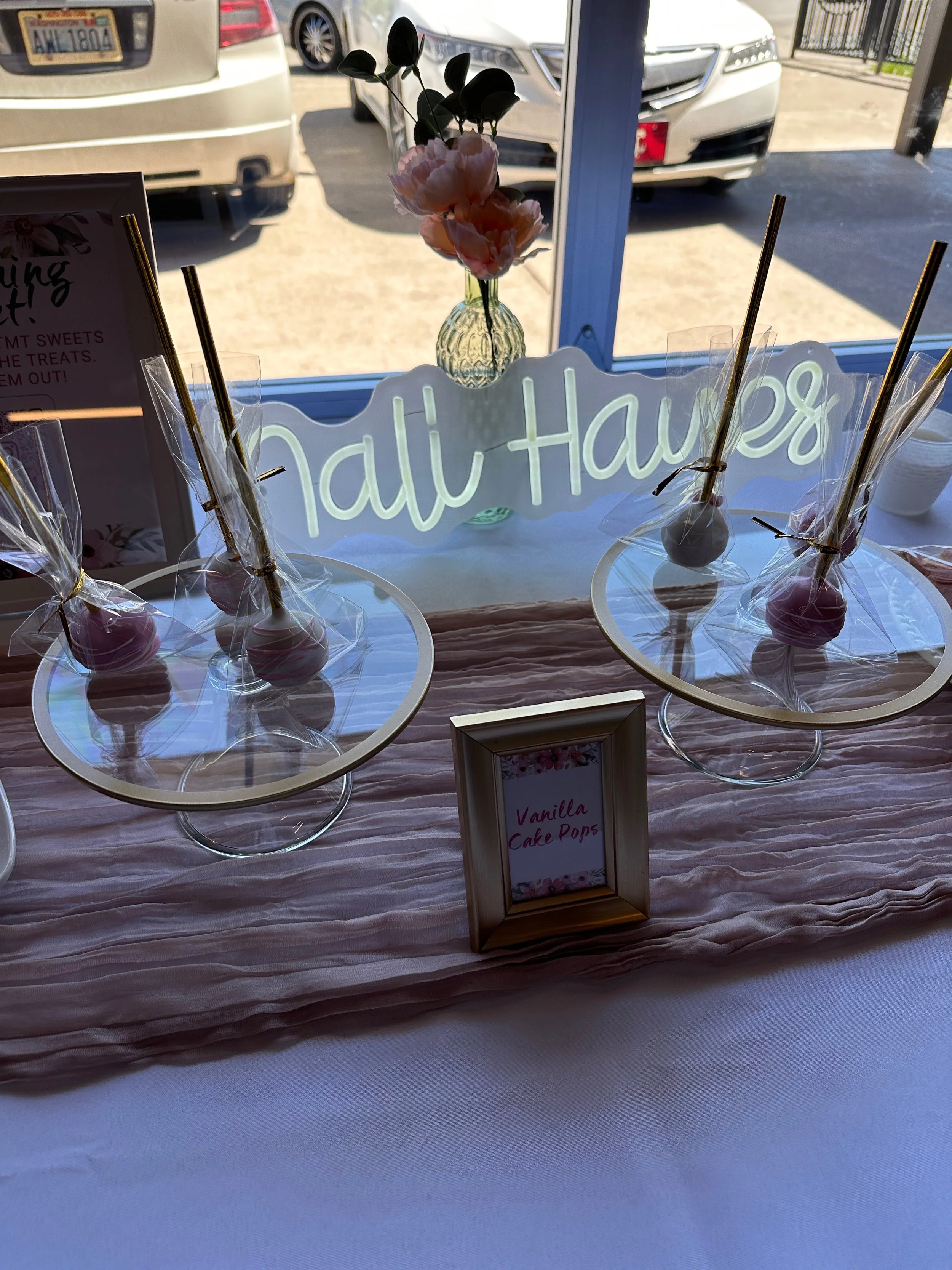 Display table with vanilla cake pops wrapped in plastic, a pink flower in a vase, a neon 'All Hues' sign, and a sign indicating 'Vanilla Cake Pops', set near a window with cars outside.