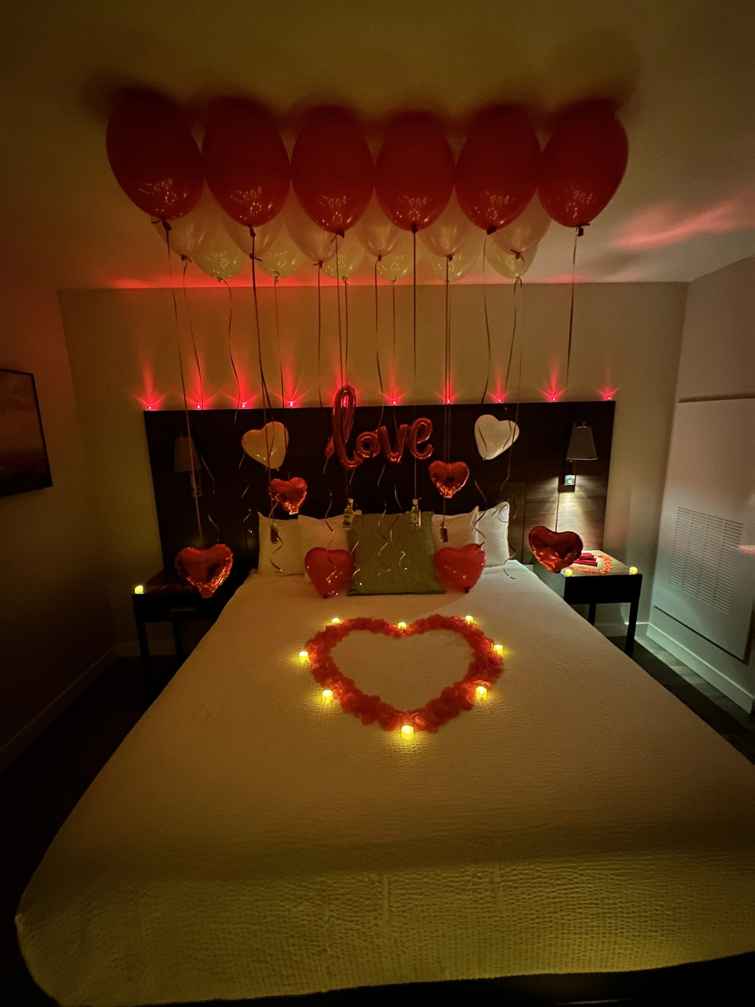Decorated hotel bed with red and white balloons floating above, illuminated heart-shaped candles, a heart made of rose petals, and themed Valentine's Day decorations.