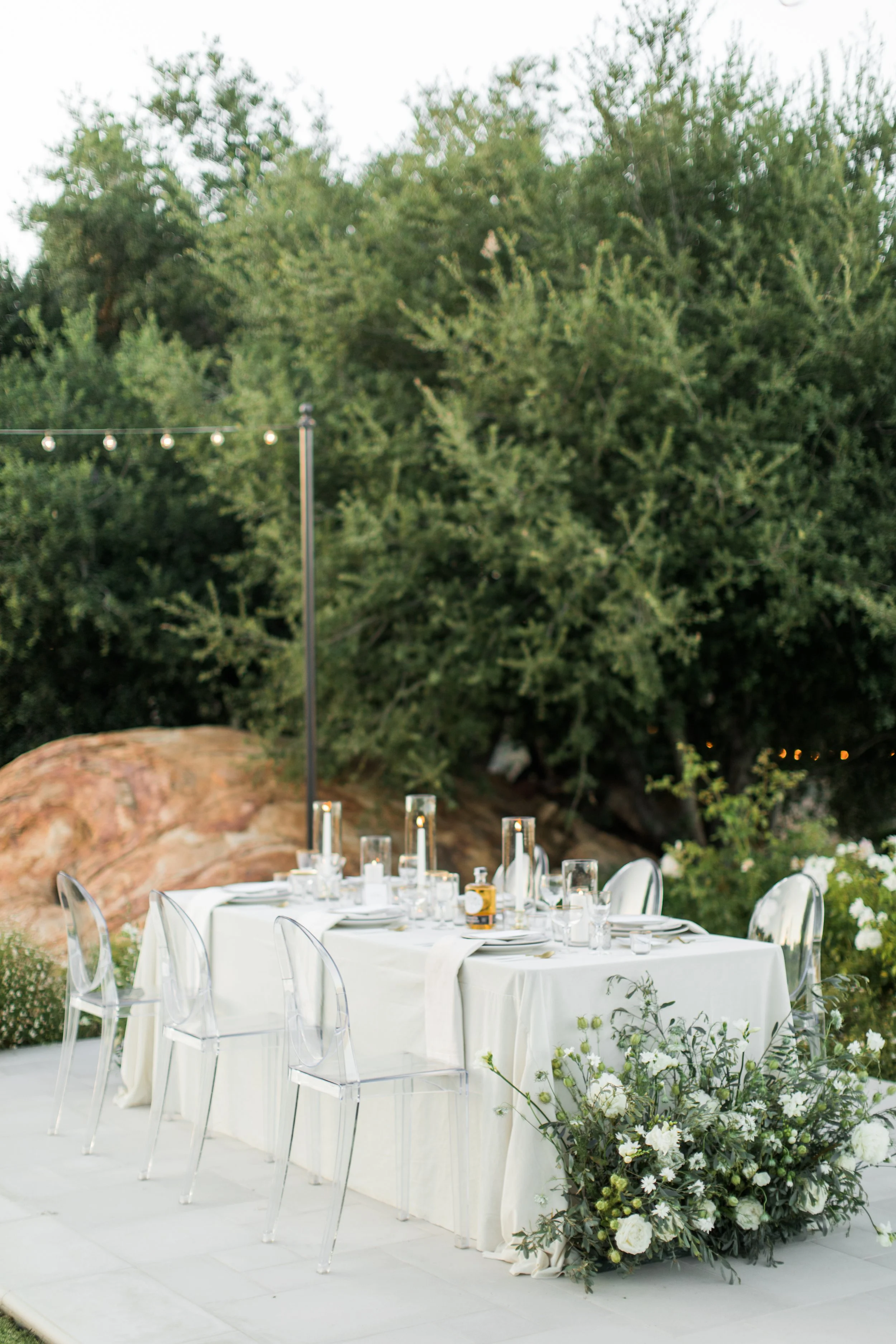 Outdoor dining table set for a celebration, decorated with white tablecloth, glassware, and floral arrangements, surrounded by greenery and rocks.