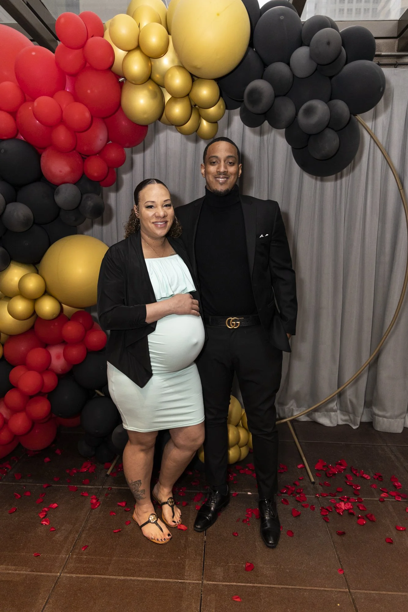 A pregnant woman and a man dressed in formal attire standing in front of a balloon arch with red, black, and gold balloons, at a celebration or event.