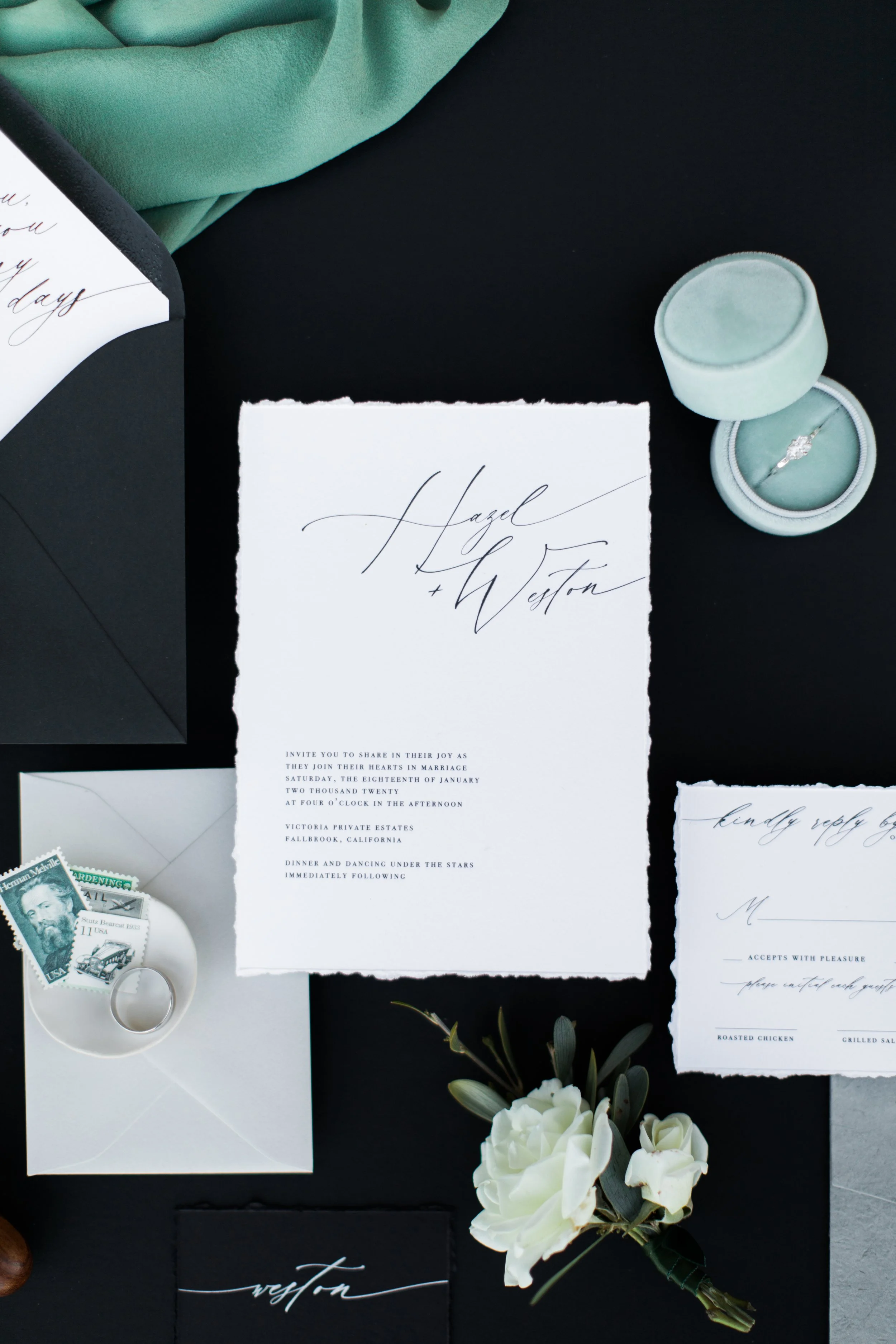 Elegant wedding invitation suite with calligraphy, vintage stamps, and a white floral boutonniere on a black surface.