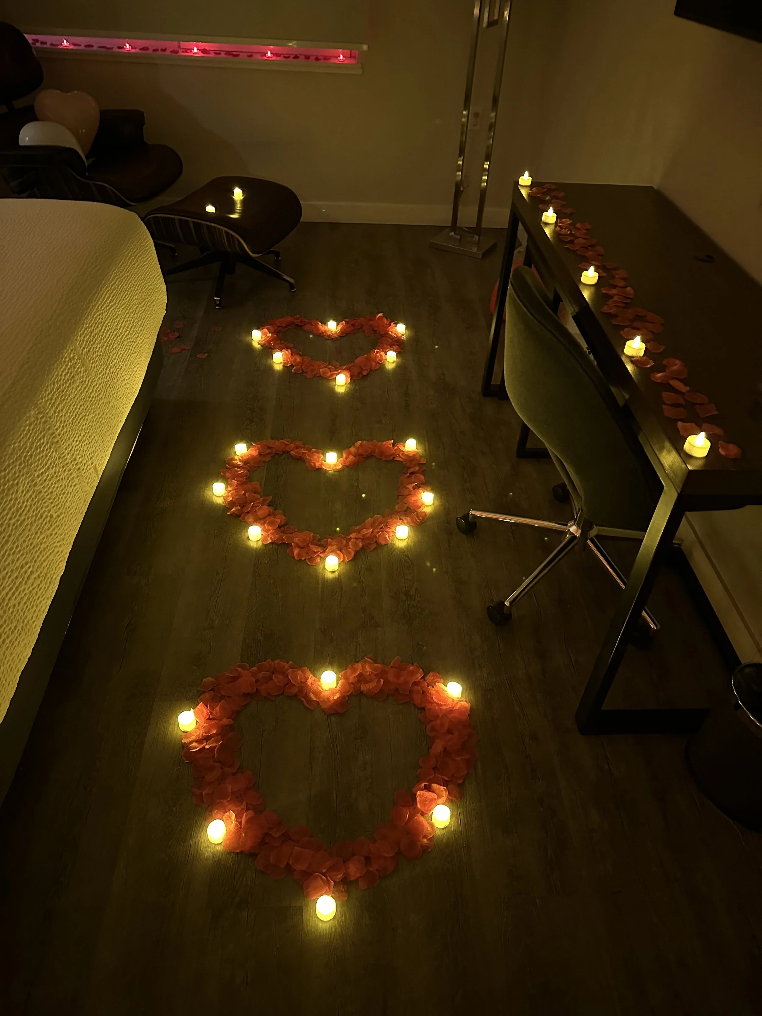 Decorative setup with heart-shaped flower arrangements outlined by small candles on a dark wood floor, creating a romantic ambiance.