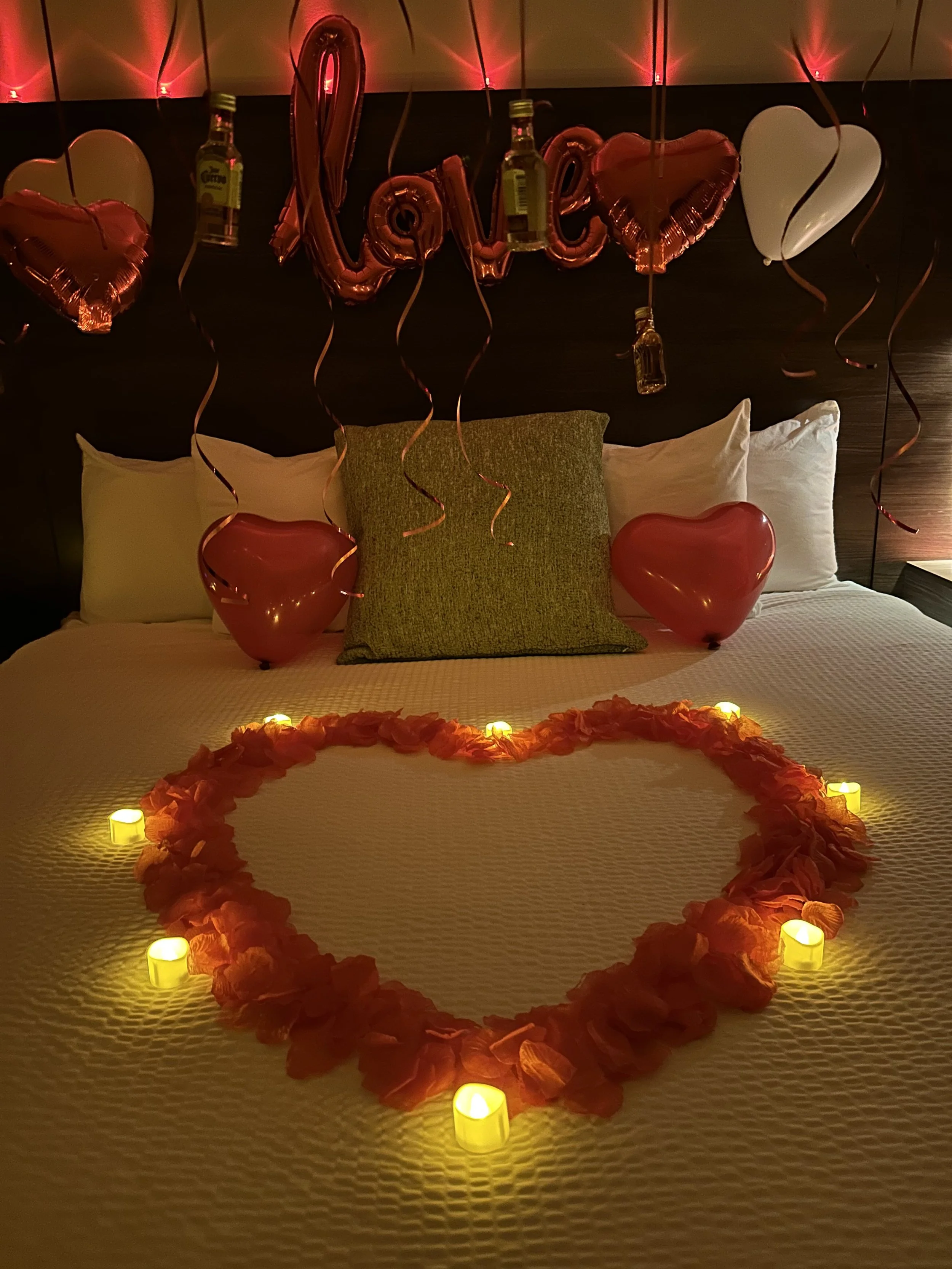 A decorated hotel bed for Valentine's Day with heart-shaped balloons, LED candles, rose petal heart, and hanging balloons spelling 'love' with a small bottle of alcohol.
