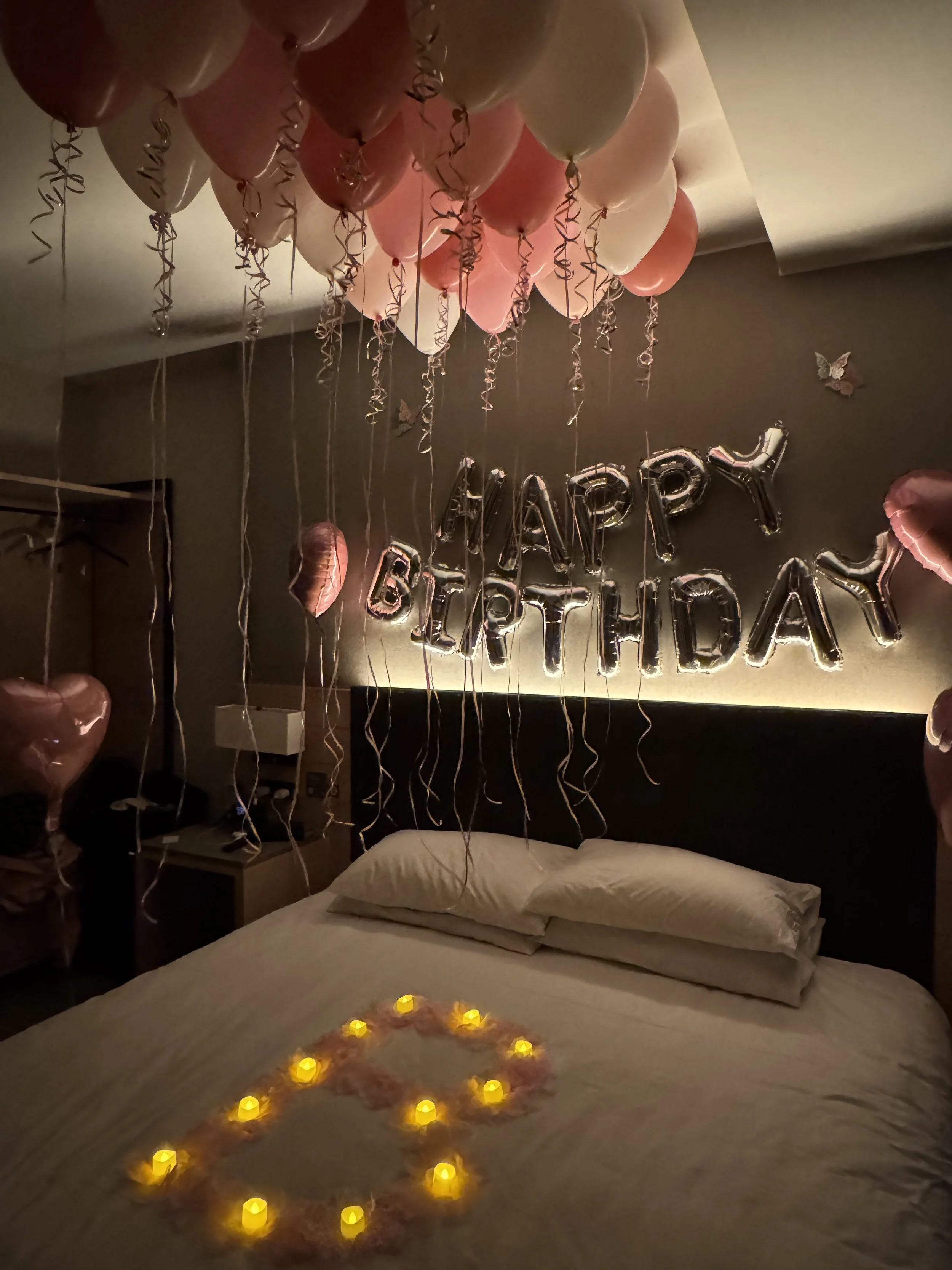 A bedroom decorated for a birthday celebration with balloons, candles, and a 'Happy Birthday' balloon banner on the wall.