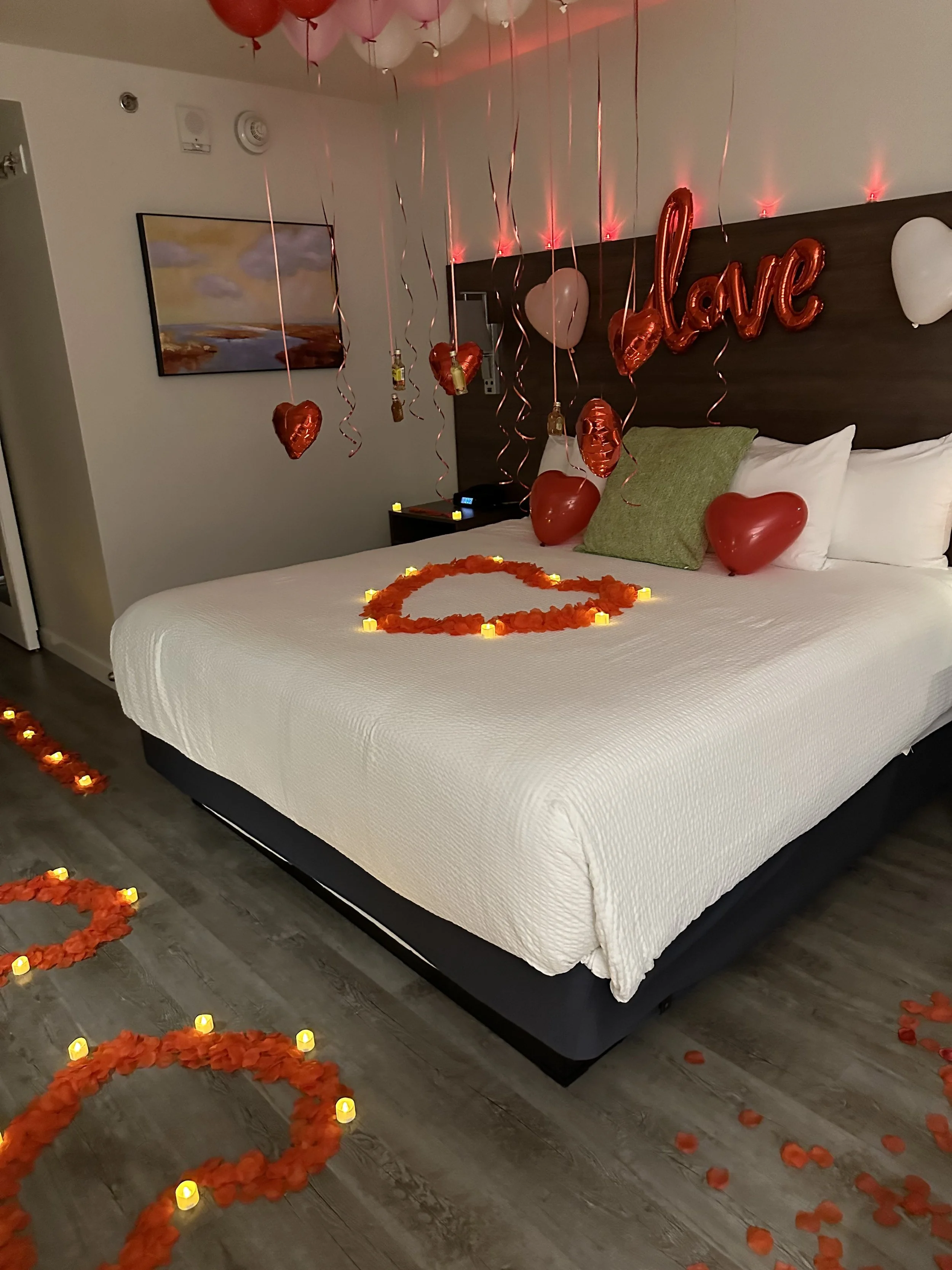 Decorated hotel bed with red heart-shaped balloons, rose petals, candles, and a 'love' balloon inside a Valentine's Day-themed setup.