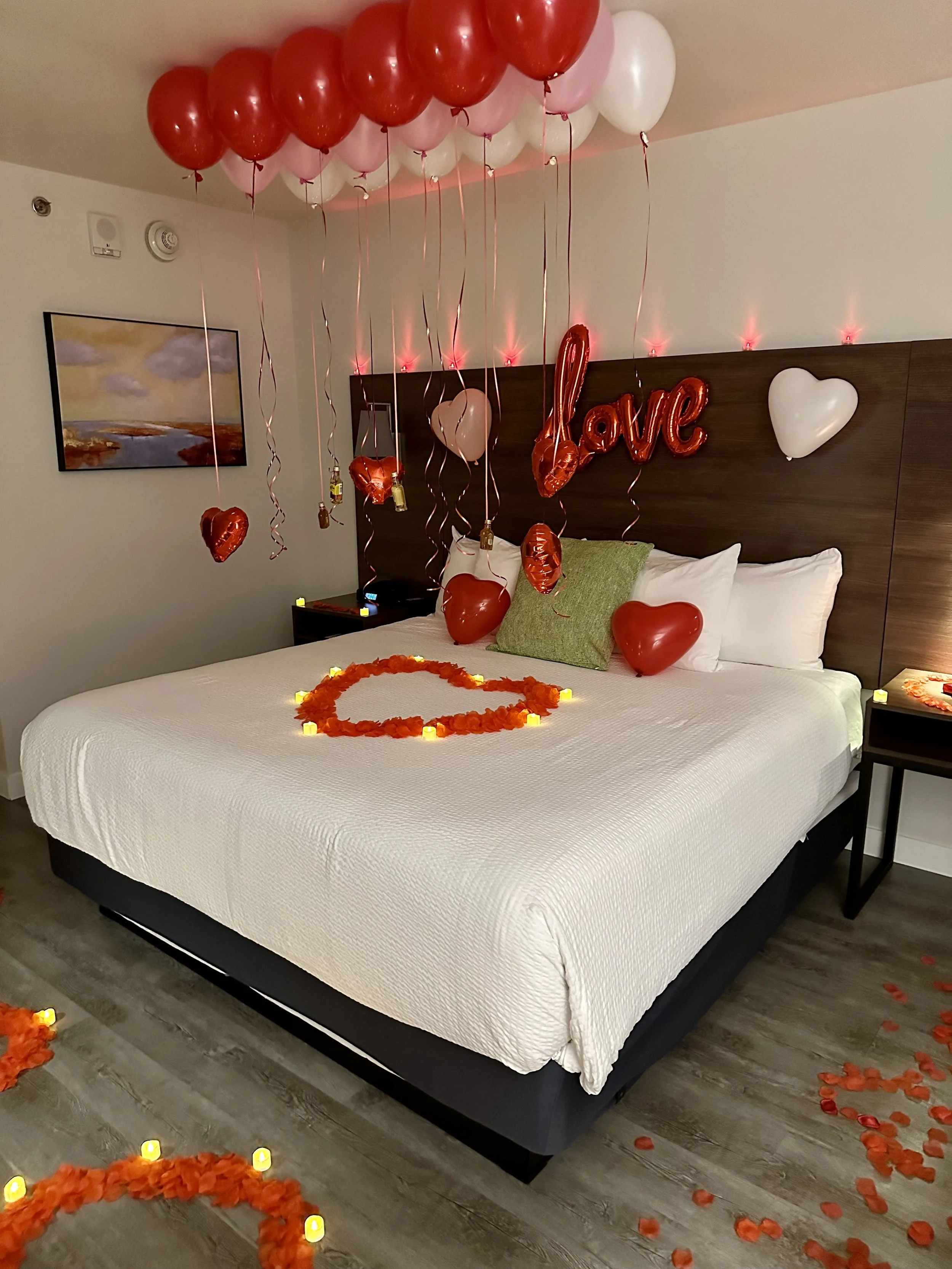 Decorated hotel room with red and pink balloons, heart-shaped balloons, a large 'love' balloon, flower petals forming a heart on the bed, and candles, suggesting a romantic celebration.