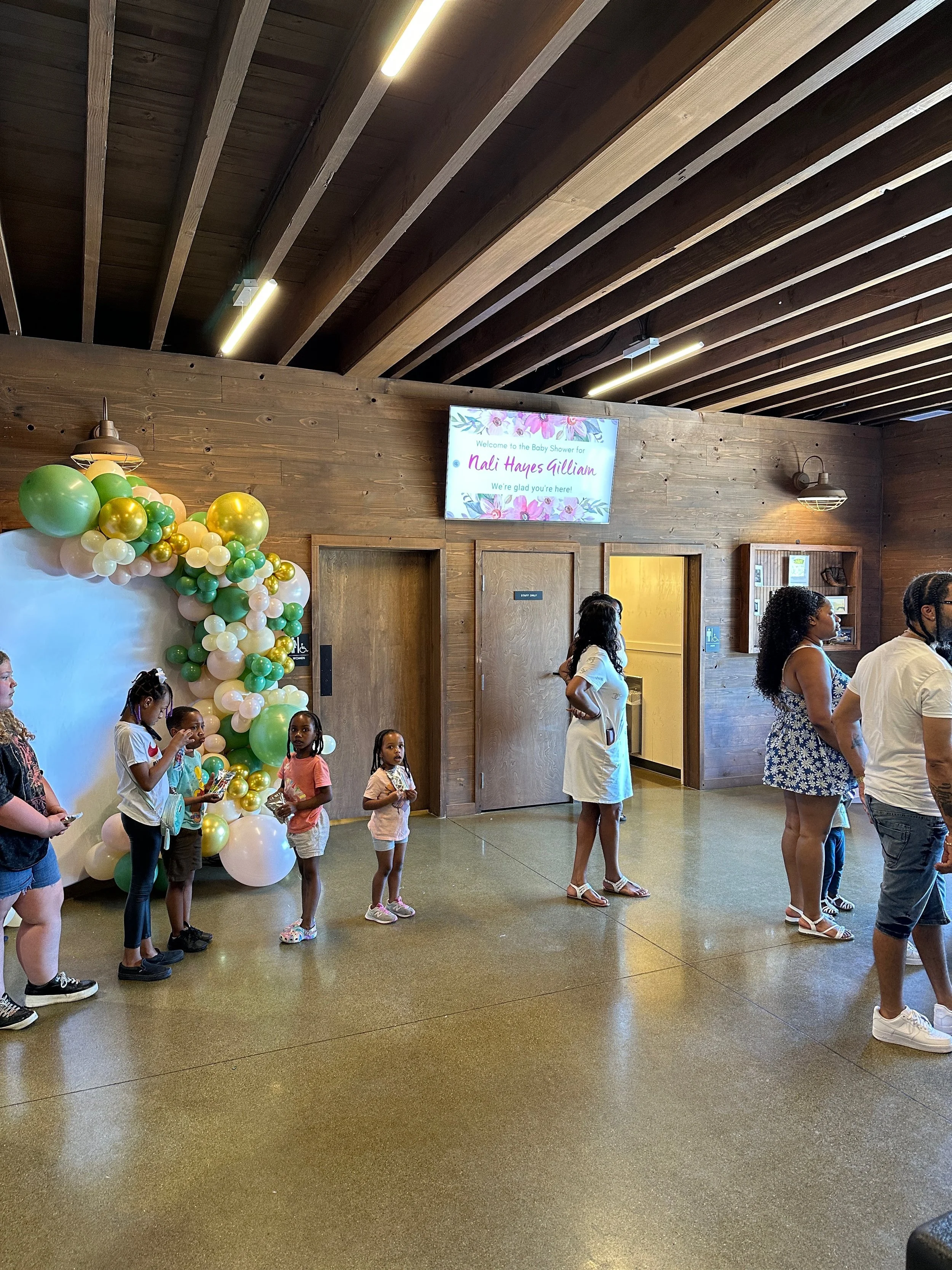 Children and adults standing in line at a baby shower decorated with balloon arch, with a digital sign welcoming guests for a baby shower for Nail Hayes Gillian showing a floral background that reads, 'Welcome to the Baby Shower for Nail Hayes Gillia