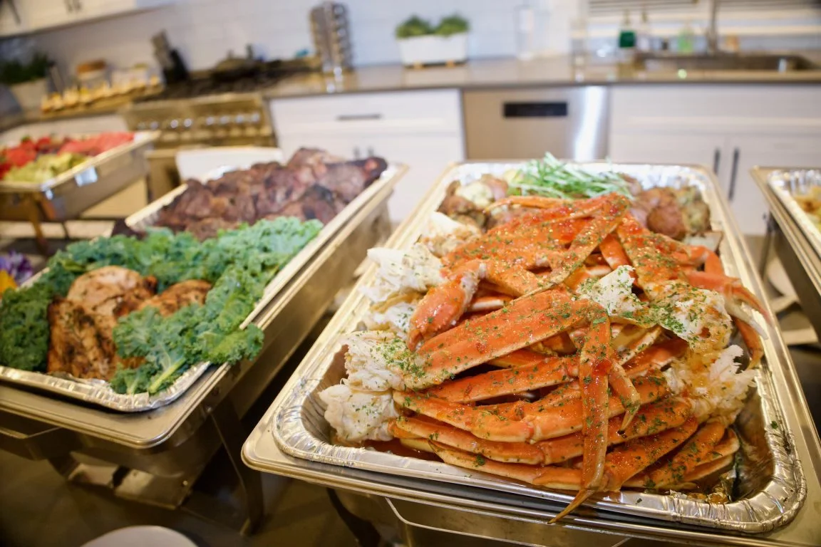 A buffet table with cooked crab legs garnished with herbs, grilled meats, and vegetables in a kitchen setting.