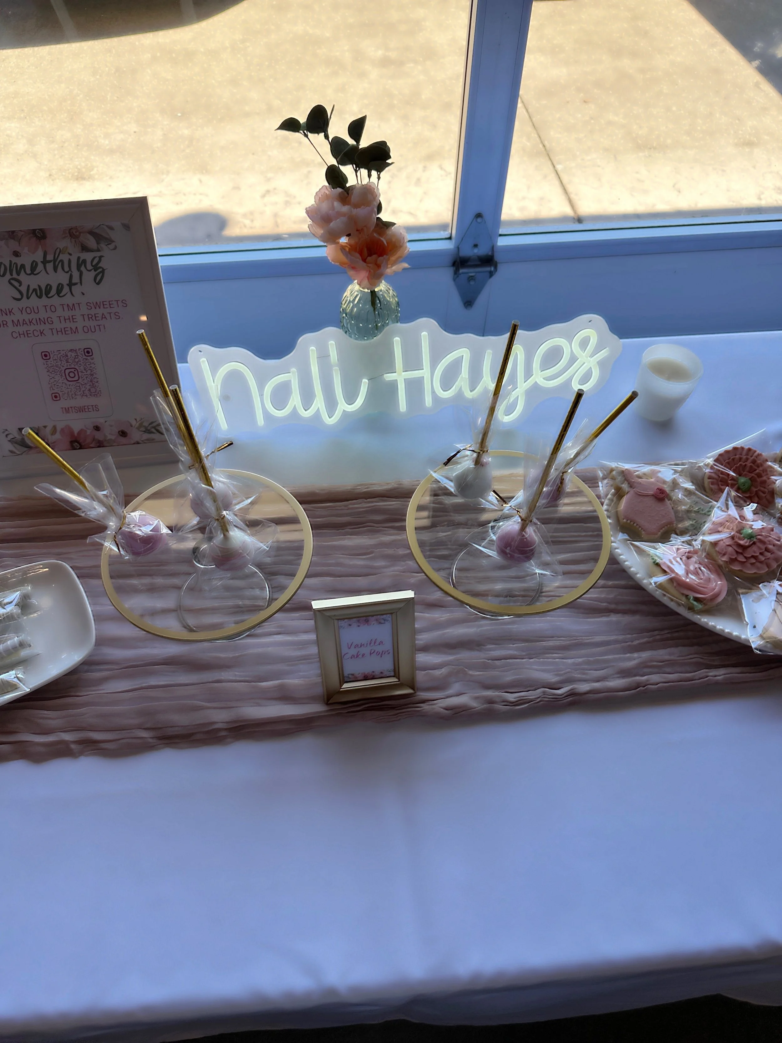 Table decorated with pink and white cookies and cake pops, a white candle, a small vase with pink flowers, a neon sign that reads 'Nail Hayes', and a sign thanking guests for making treats, at a sweet-themed event.