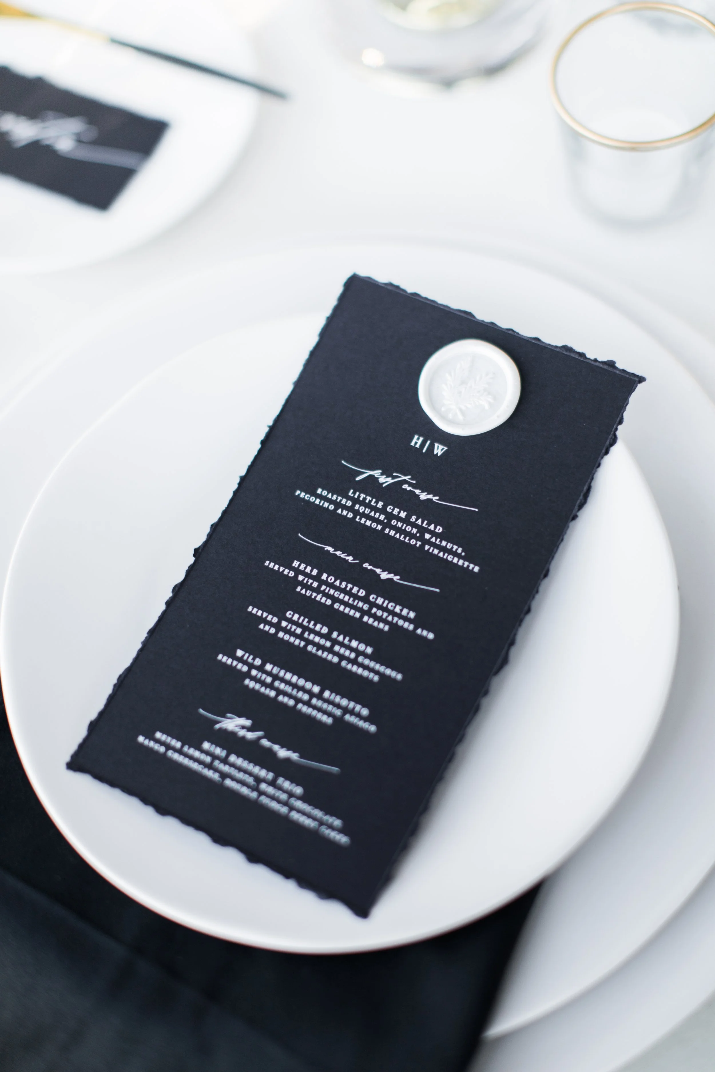 A black menu card with white text and a white wax seal on top, placed on a white plate at a formal dining setting with glasses and utensils in the background.
