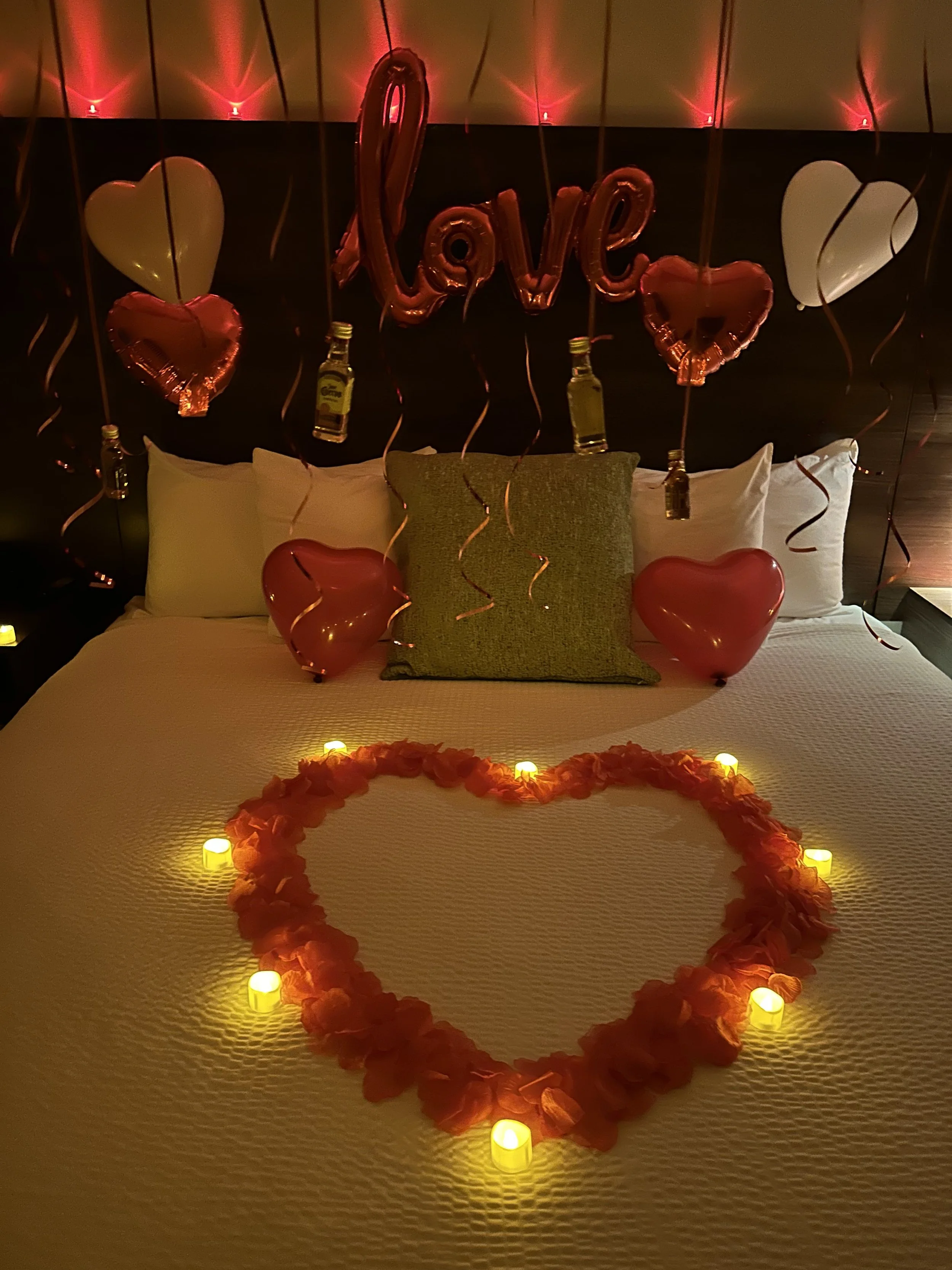 Decorated bed with heart-shaped balloons, candles, and overhead balloons spelling 'love' for romantic celebration.