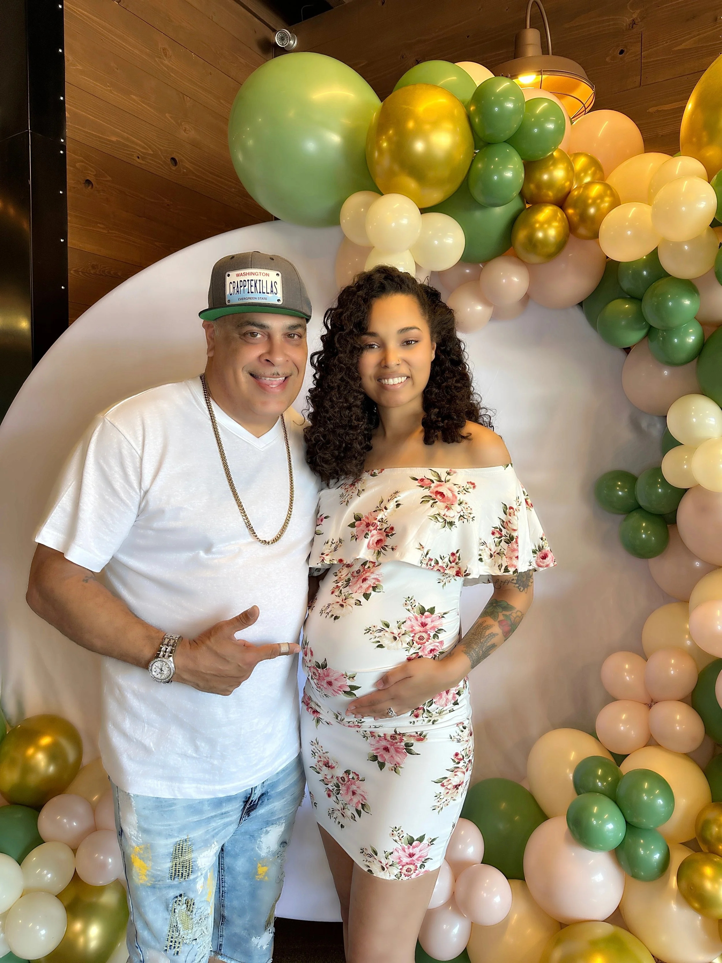A pregnant woman in a floral dress standing next to a man wearing a white t-shirt, ripped jeans, and a cap, in front of a balloon arch decoration with green, gold, and cream-colored balloons.