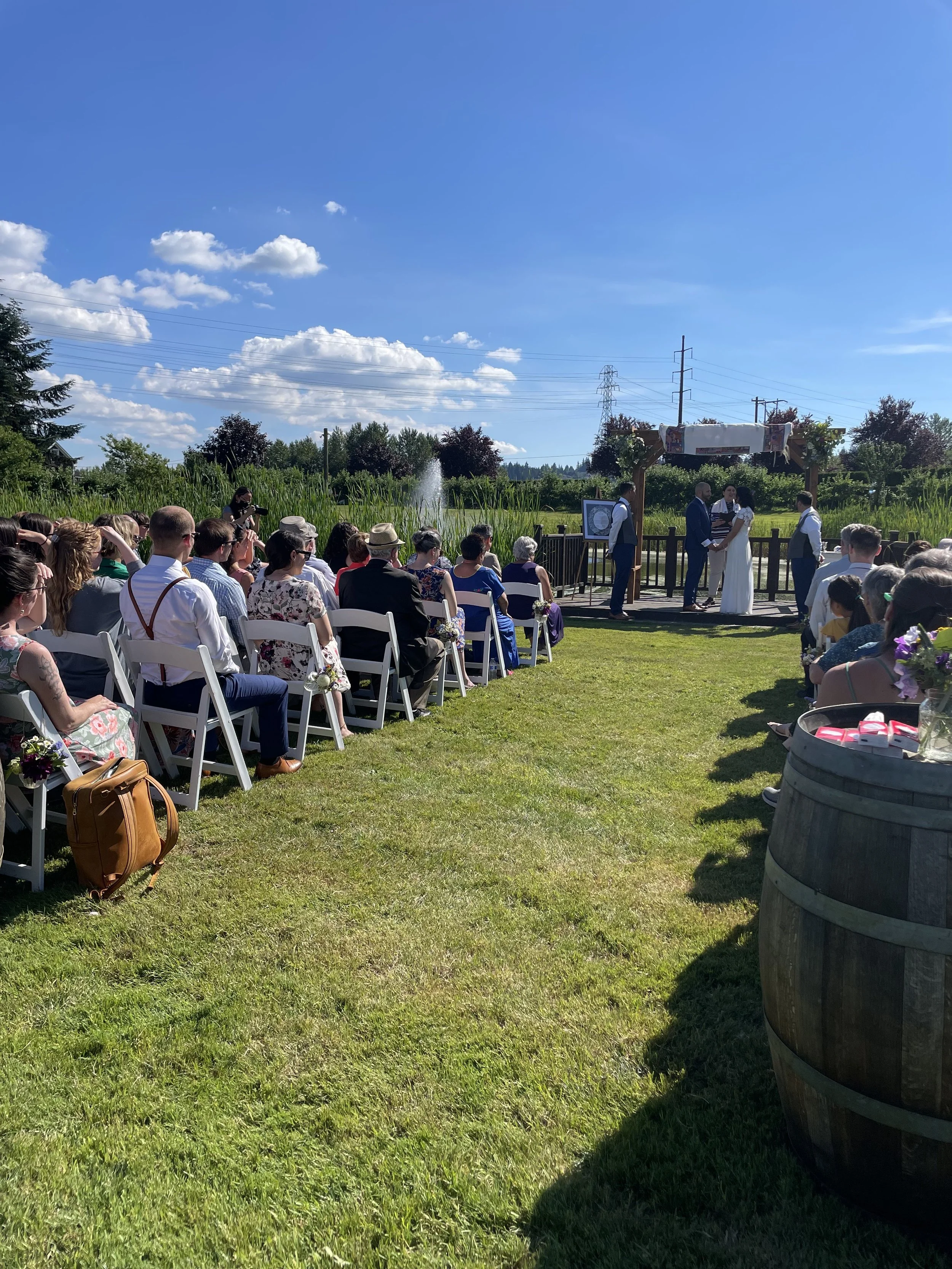 Outdoor wedding ceremony with guests seated on white chairs, officiant and bride and groom exchanging vows on a wooden platform under a decorated arch, lush green landscape, blue sky with clouds.