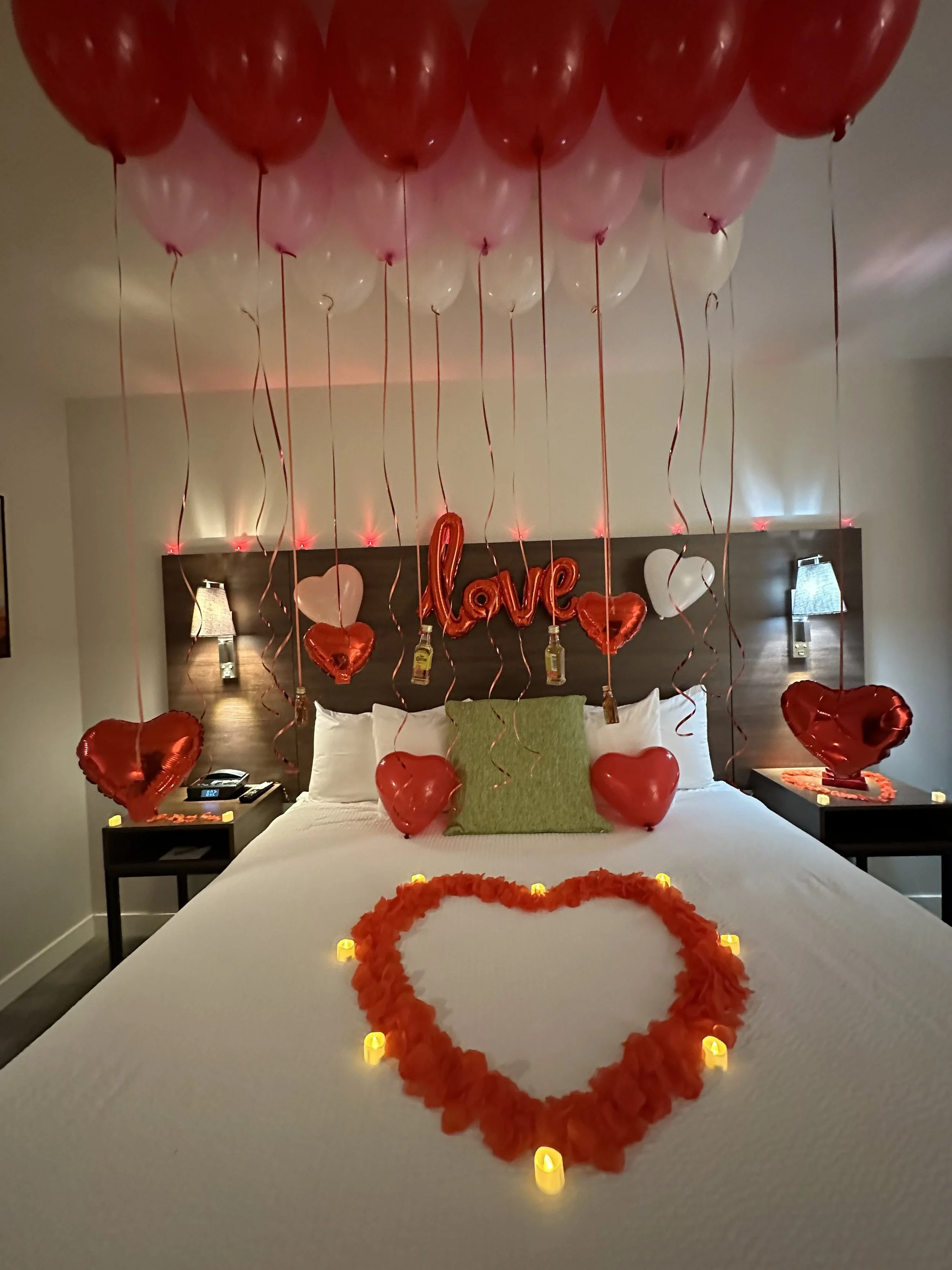Decorated hotel bed with red and pink balloons, heart-shaped balloons, candles, and Valentine's Day decorations including a heart made of flower petals on the bed and a red hearts across the headboard with a "love" sign.
