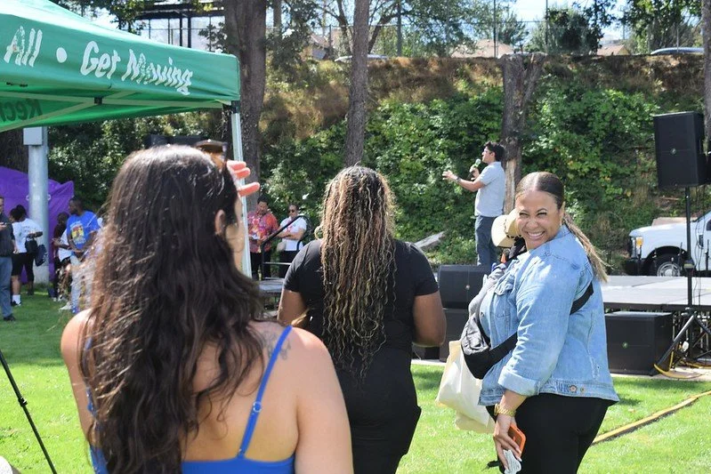 People attending an outdoor event with a stage and sound system, under a green canopy that reads 'Get Moving.' The scene includes a woman smiling at the camera, a group of people facing the stage, and trees in the background.