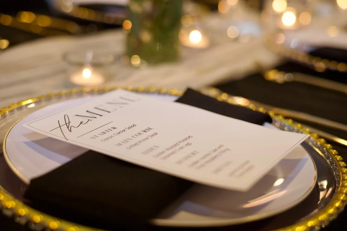 Elegant table setting with a thank you card on a plate, black napkin, and gold decorative accents, blurred background with warm lighting