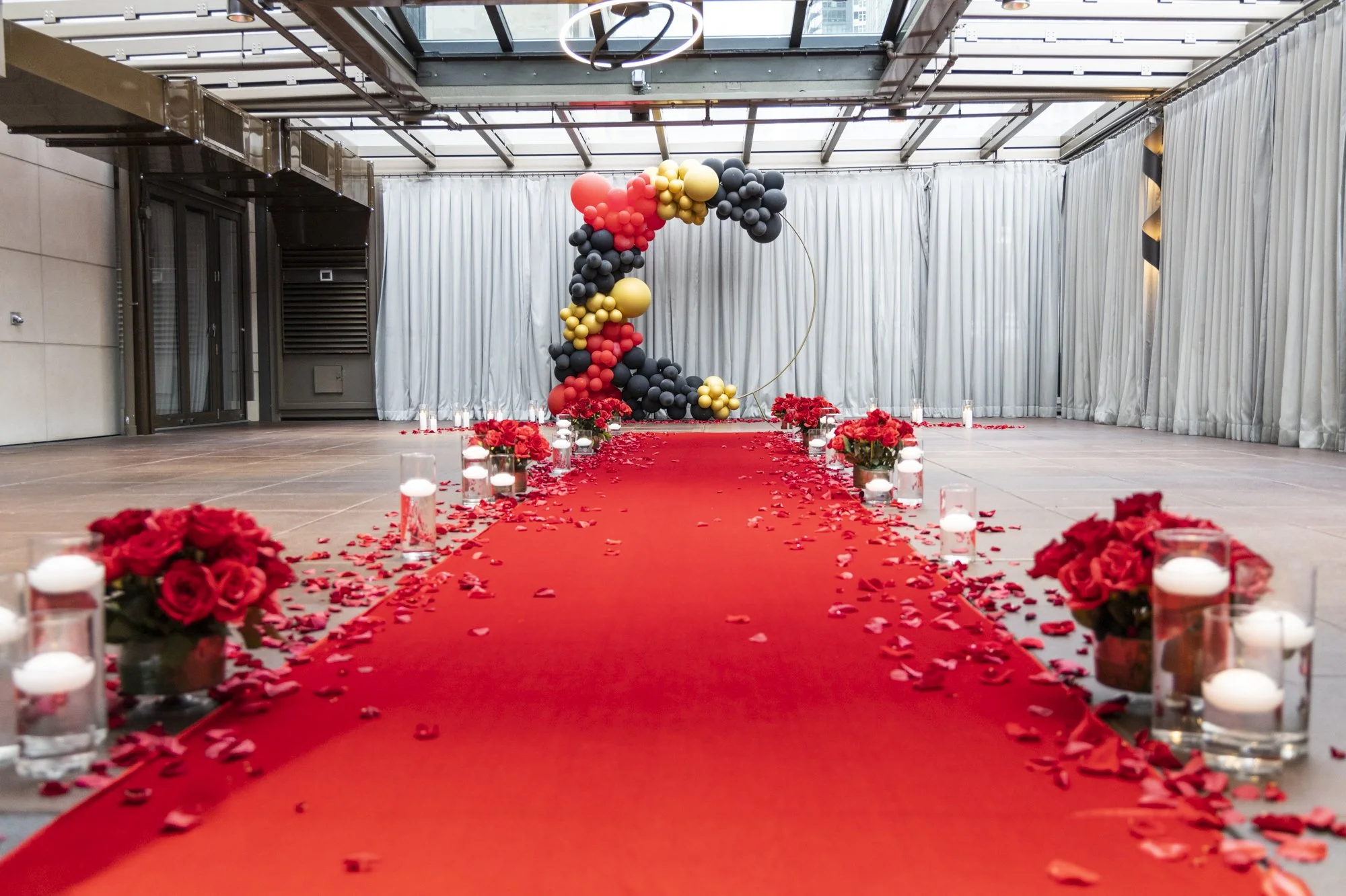 Red carpet aisle decorated with rose petals, candles, and flower arrangements on either side leading to a backdrop with balloons in red, black, and gold in an event space.