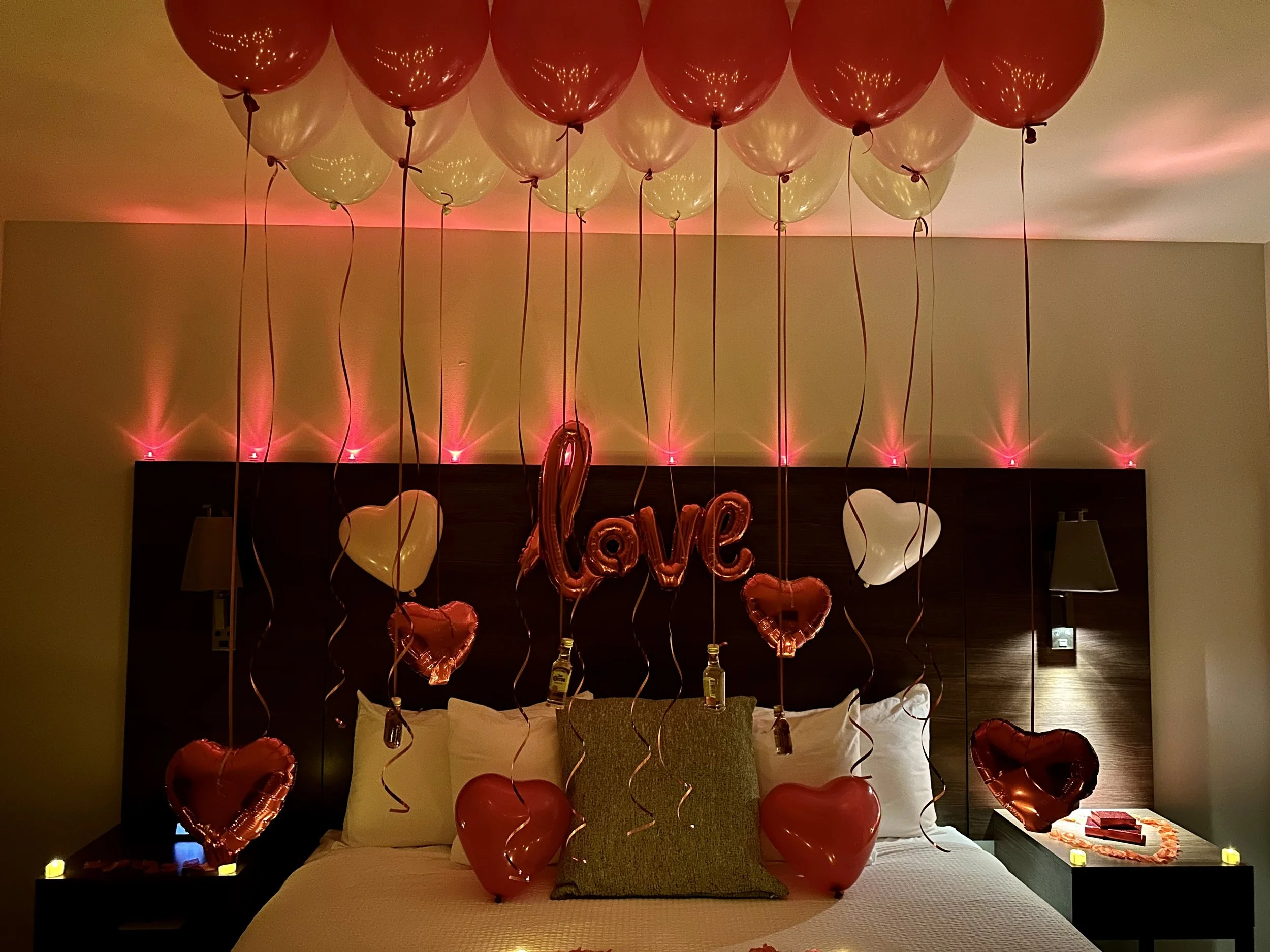 Decorated hotel room with red and white balloons, hearts, and a balloon spelling out "love" hanging above the bed for a romantic occasion.
