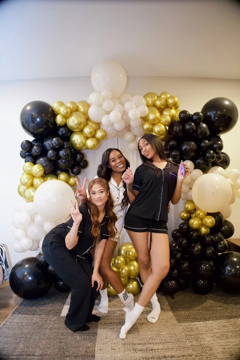 Three women in pajamas posing with peace signs in front of a balloon arch made of white, gold, and black balloons.