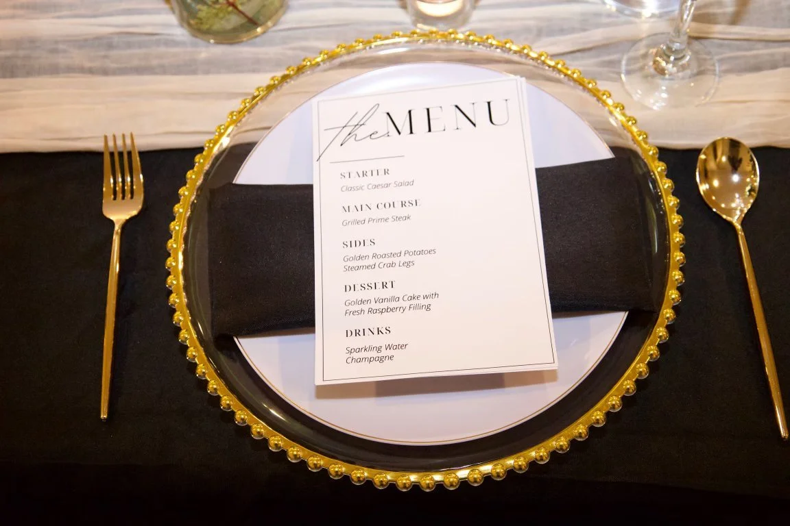 Elegant table setting with a black tablecloth, gold cutlery, a decorative gold beaded charger plate, a white dinner plate, and a menu placed on a black cloth napkin. The menu lists a starter, main course, sides, dessert, and drinks.