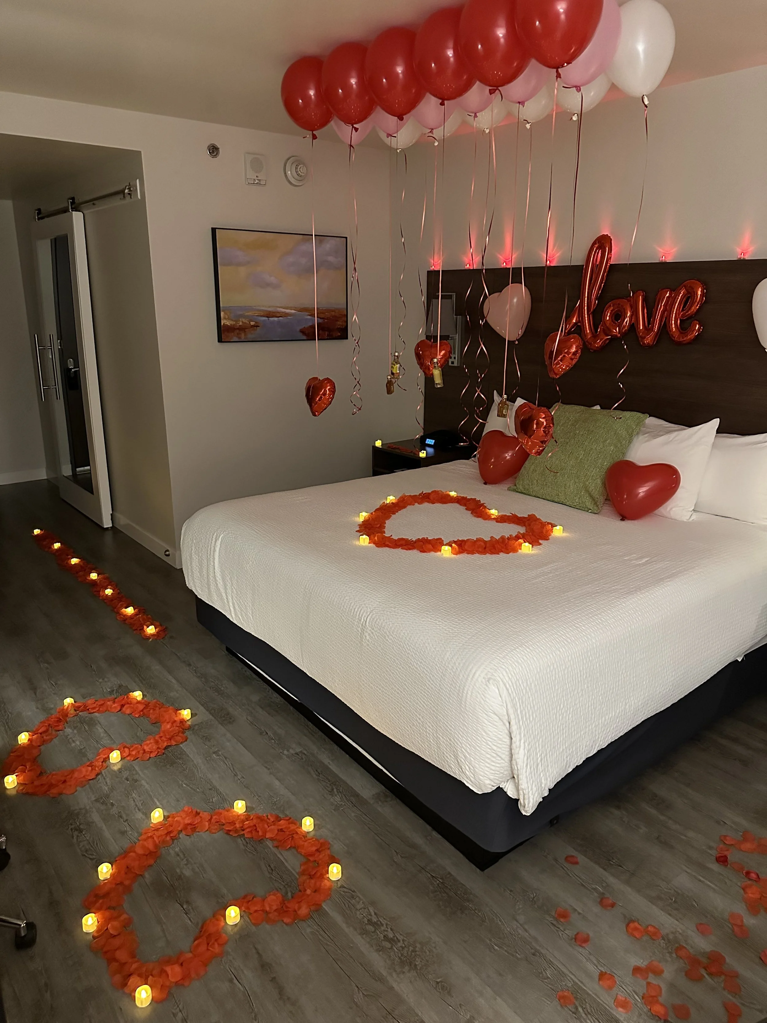 Decorated hotel room for Valentine's Day with red, pink, and white balloons, heart-shaped floral arrangements, LED candles, and a 'love' balloon sign on the headboard.