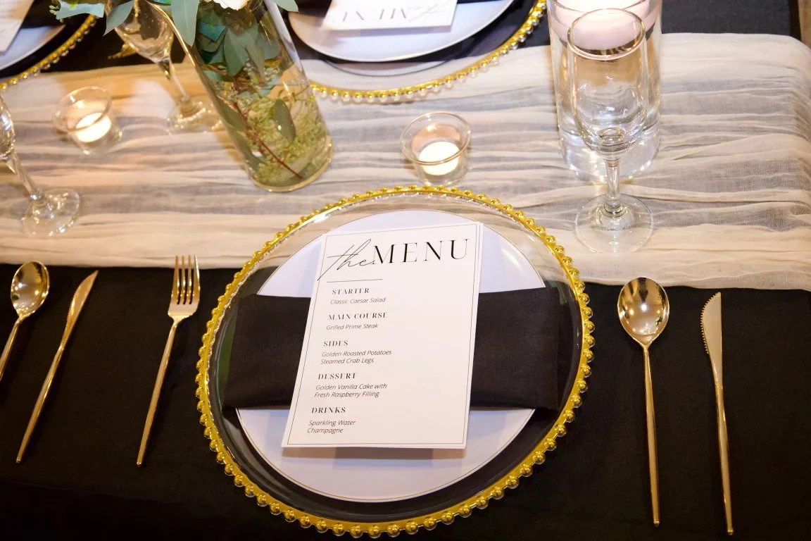 Elegant dining table with gold and black accents, featuring a place setting with a menu on a black napkin, gold utensils, and decorative elements like a floral centerpiece and candles.