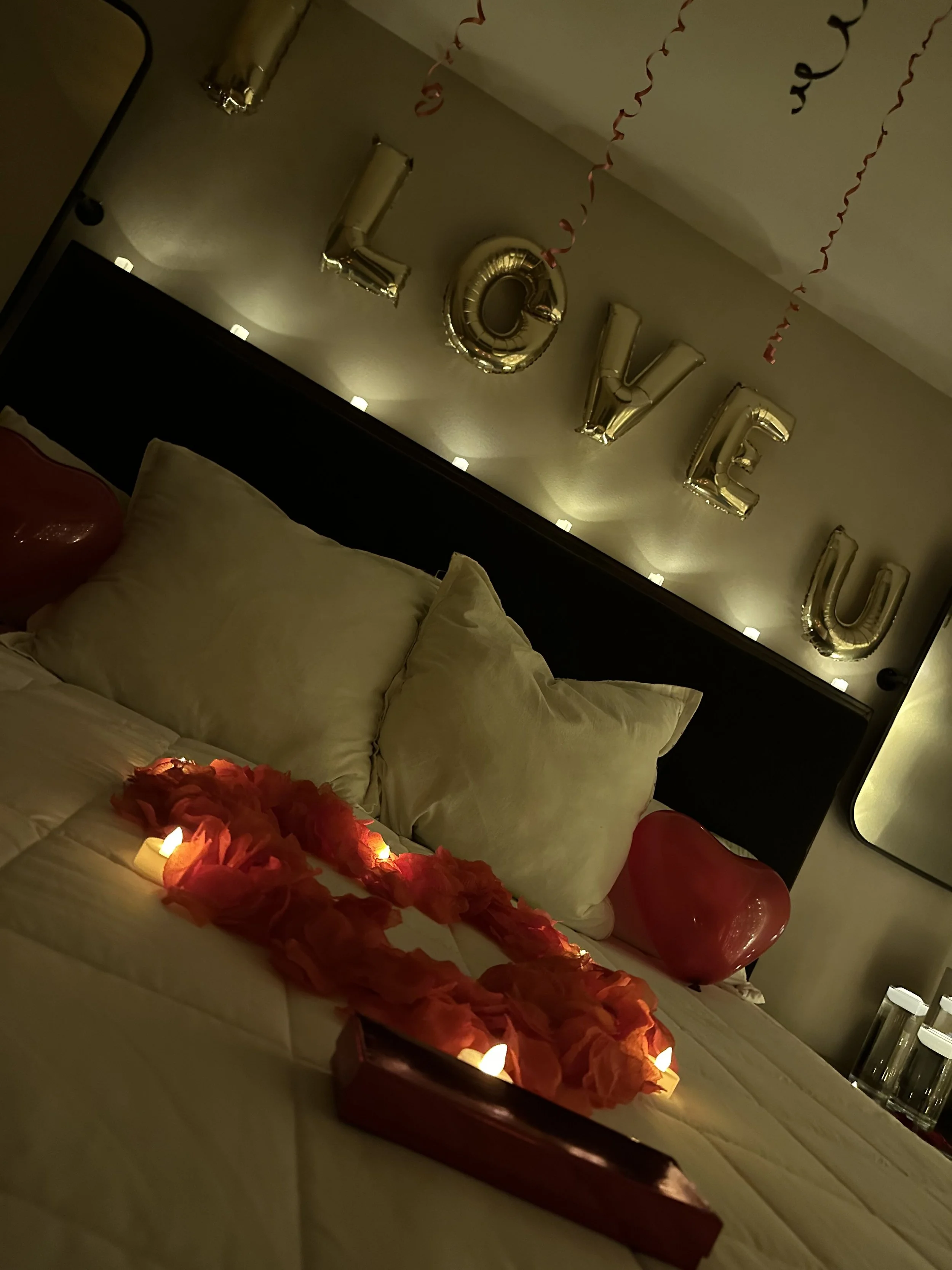 A decorated bed with heart-shaped balloons, candles, and flower petals arranged in a heart shape, with a 