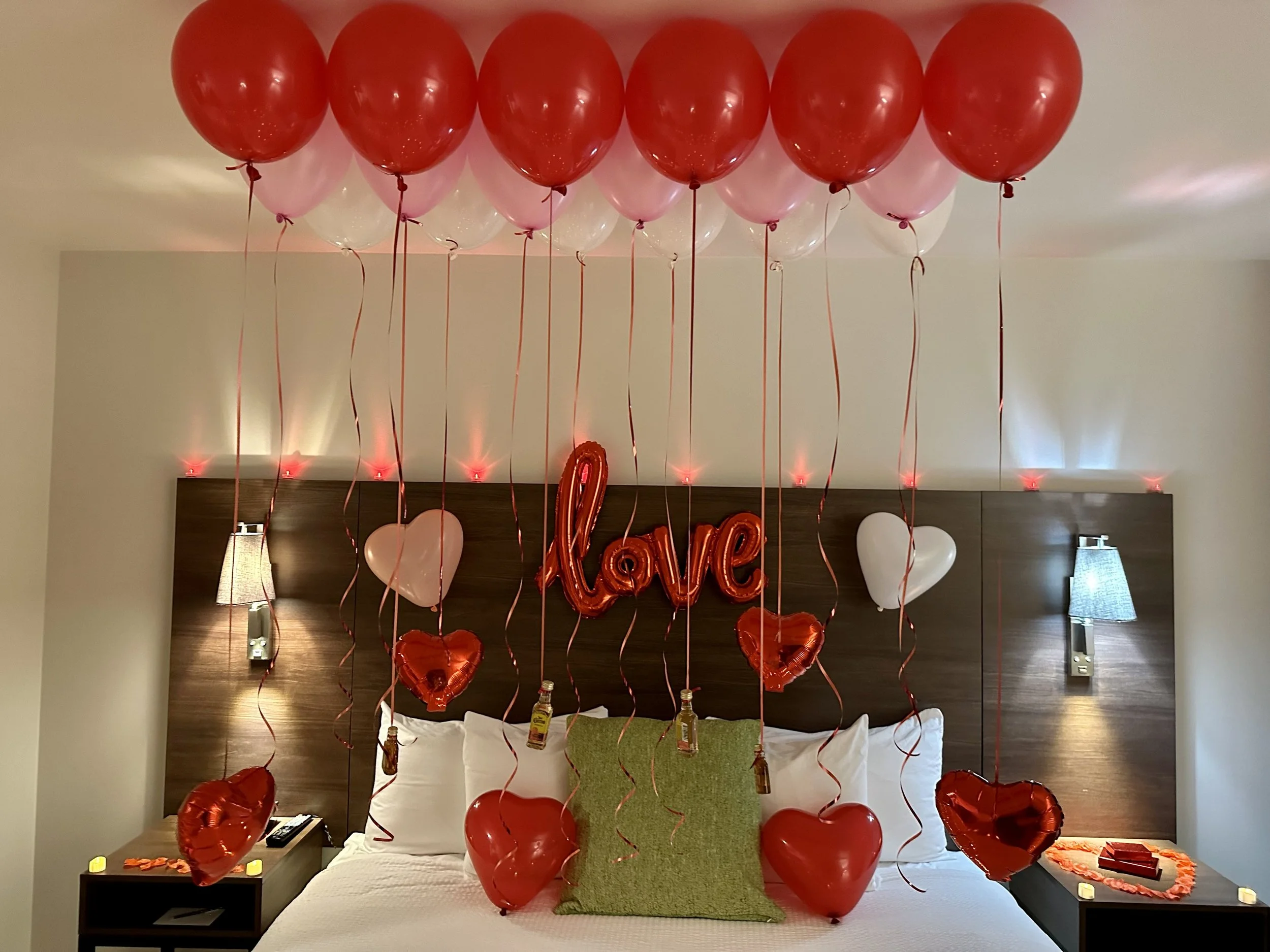 Valentine's Day decorated hotel room with red and pink balloons hanging from the ceiling, heart-shaped balloons on the bed, and a 'Love' sign on the headboard.