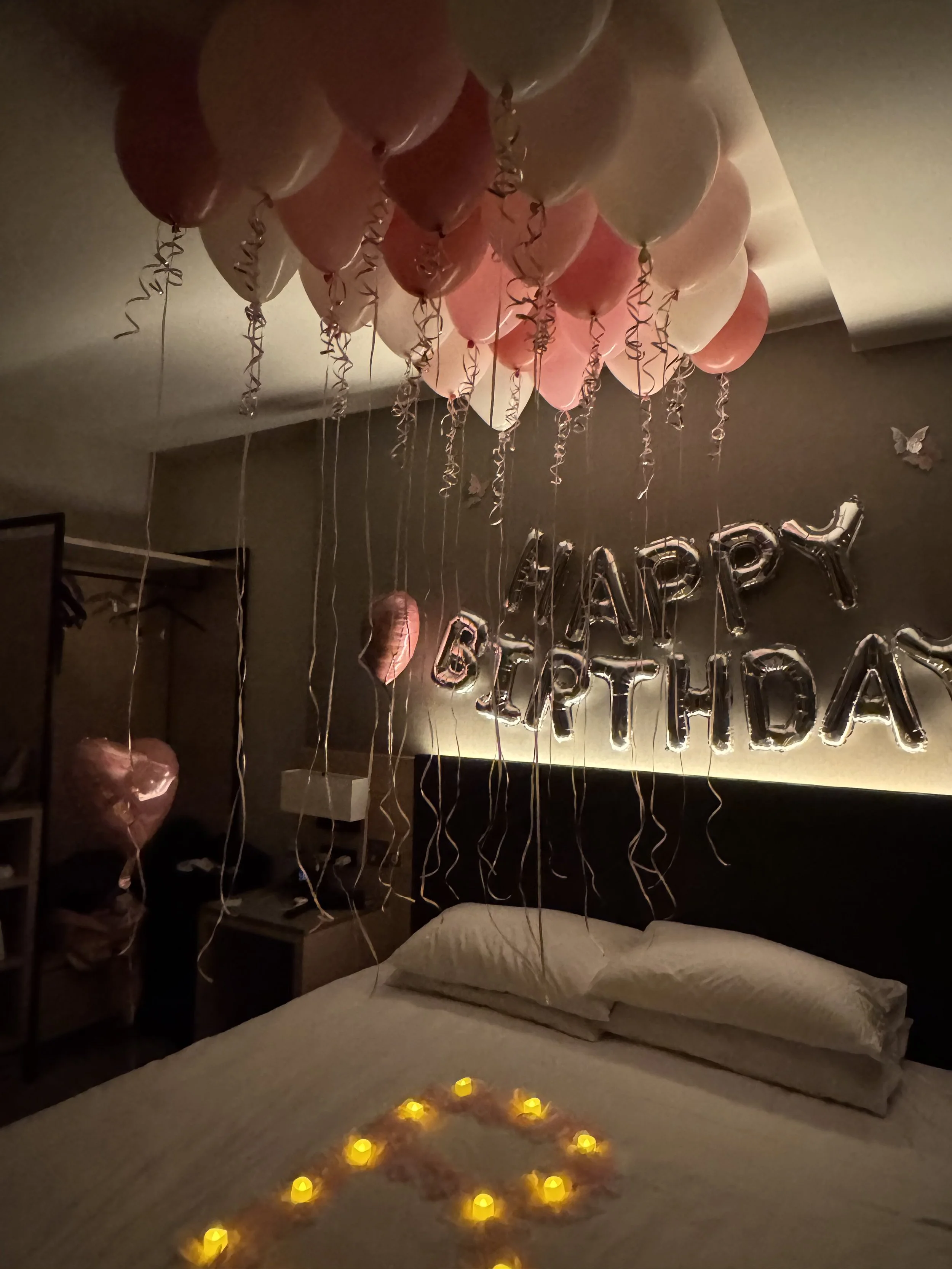 Decorated bedroom with pink, white, and peach balloons with curly ribbons, a 'Happy Birthday' balloon banner on the wall, a heart-shaped balloon on the bed, and lit candles arranged in the shape of a heart on the bed.