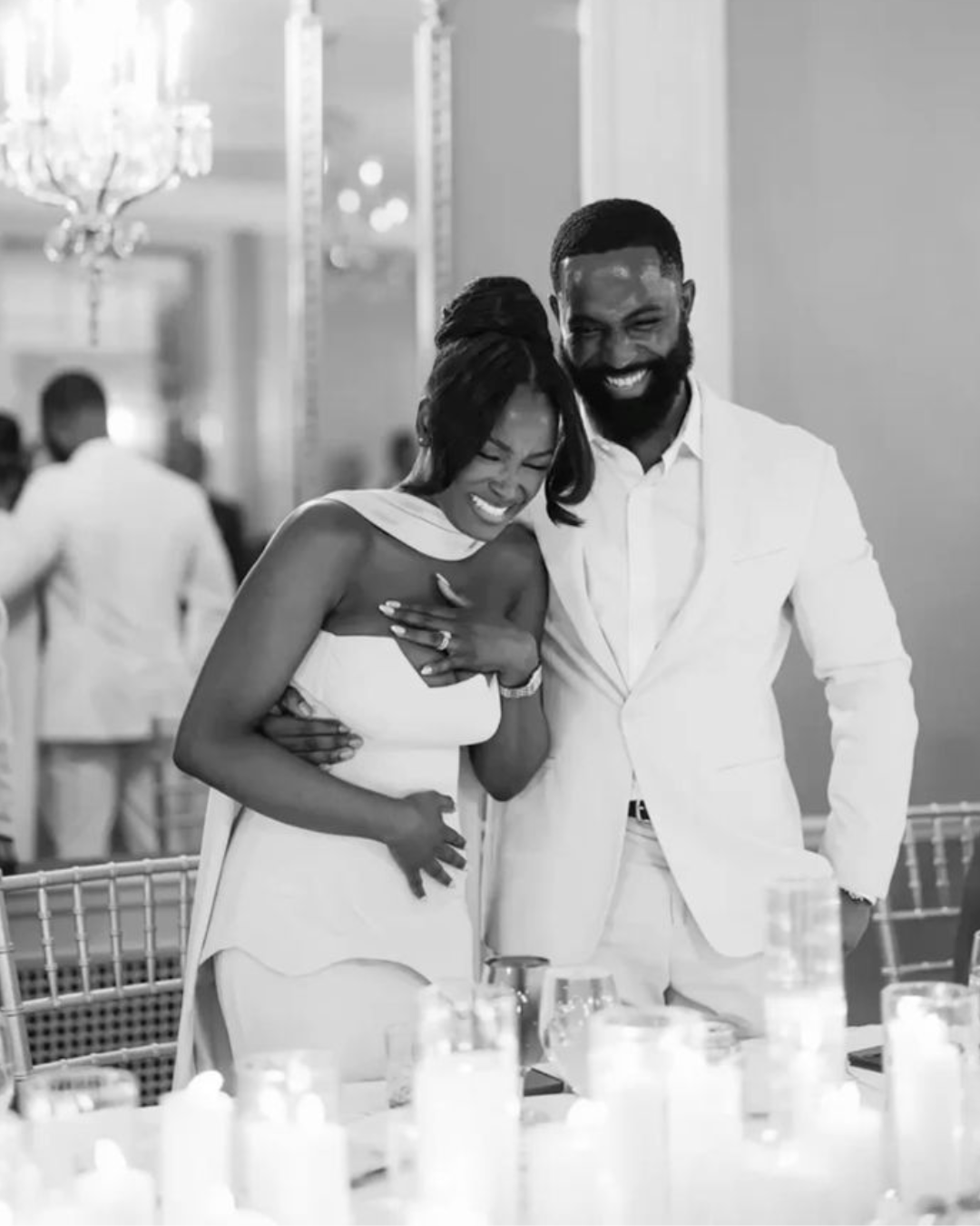 Two people laughing and embracing at a formal event, with candles and glasses on a table in the foreground.