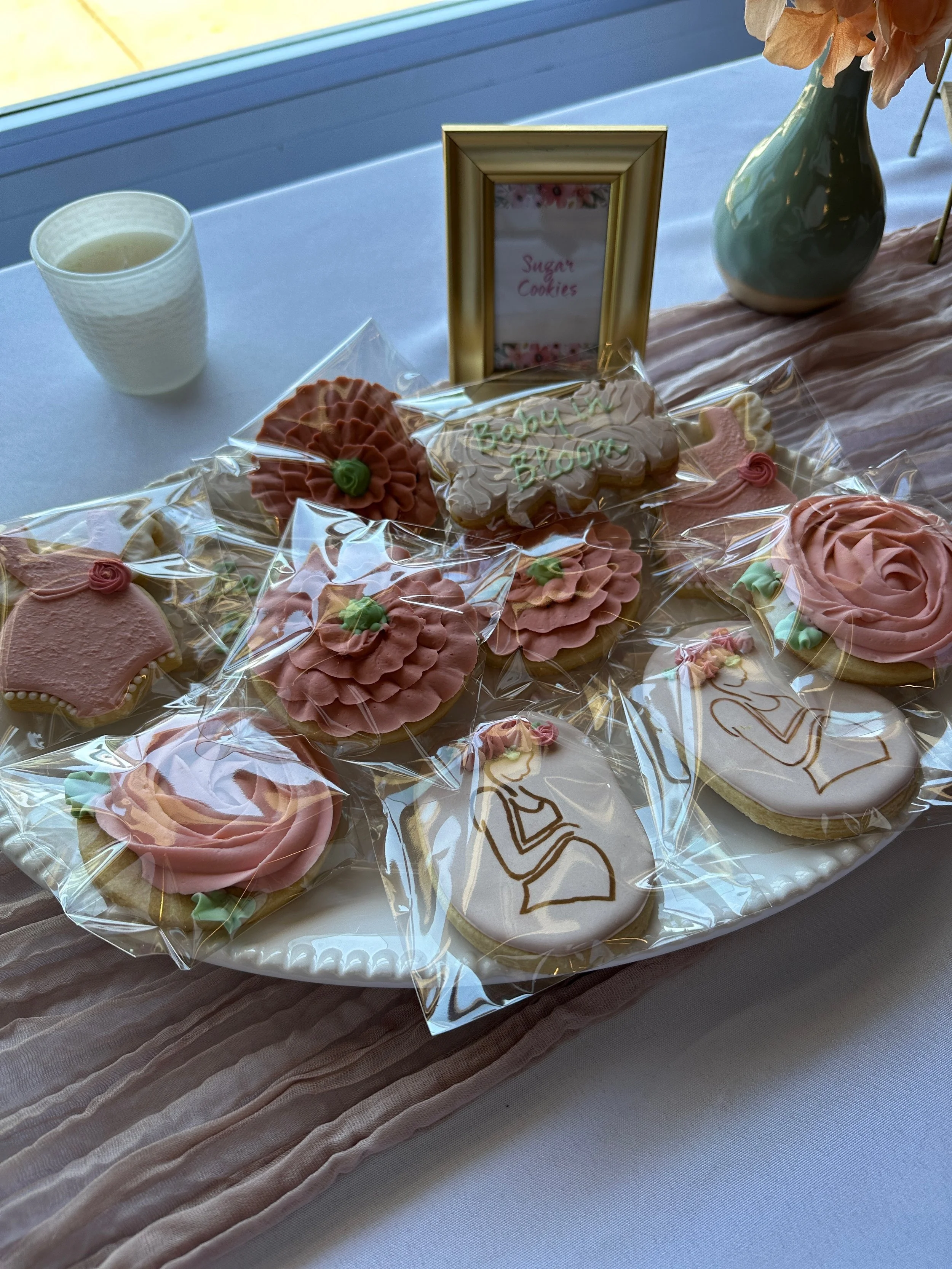 Sugar cookies decorated with pink, white, and green icing, some with floral and abstract designs, arranged on a white platter with clear wrapping, on a table with a beige table runner, a gold picture frame labeled 'Sugar Cookies', a white cup, and a 