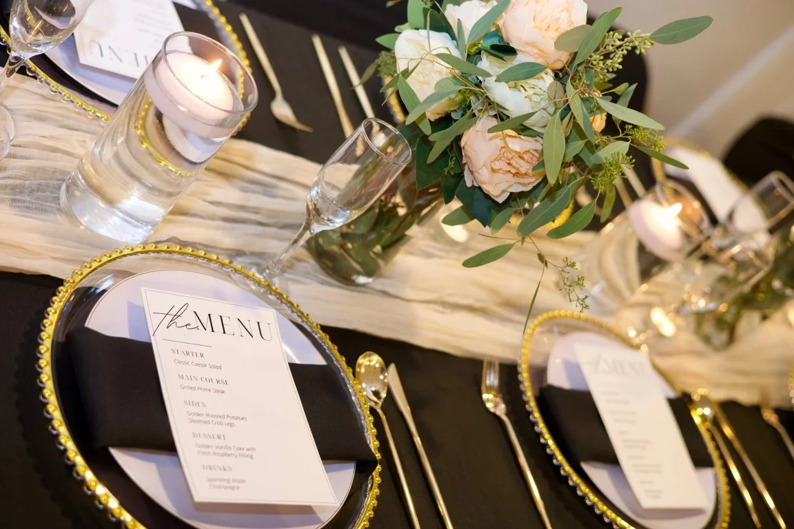Elegant banquet table setting with a black tablecloth, gold-rimmed charger plates, black napkins, and printed menus. Centerpieces include blush and white peonies with greenery in a glass vase, surrounded by lit candles in glass holders.