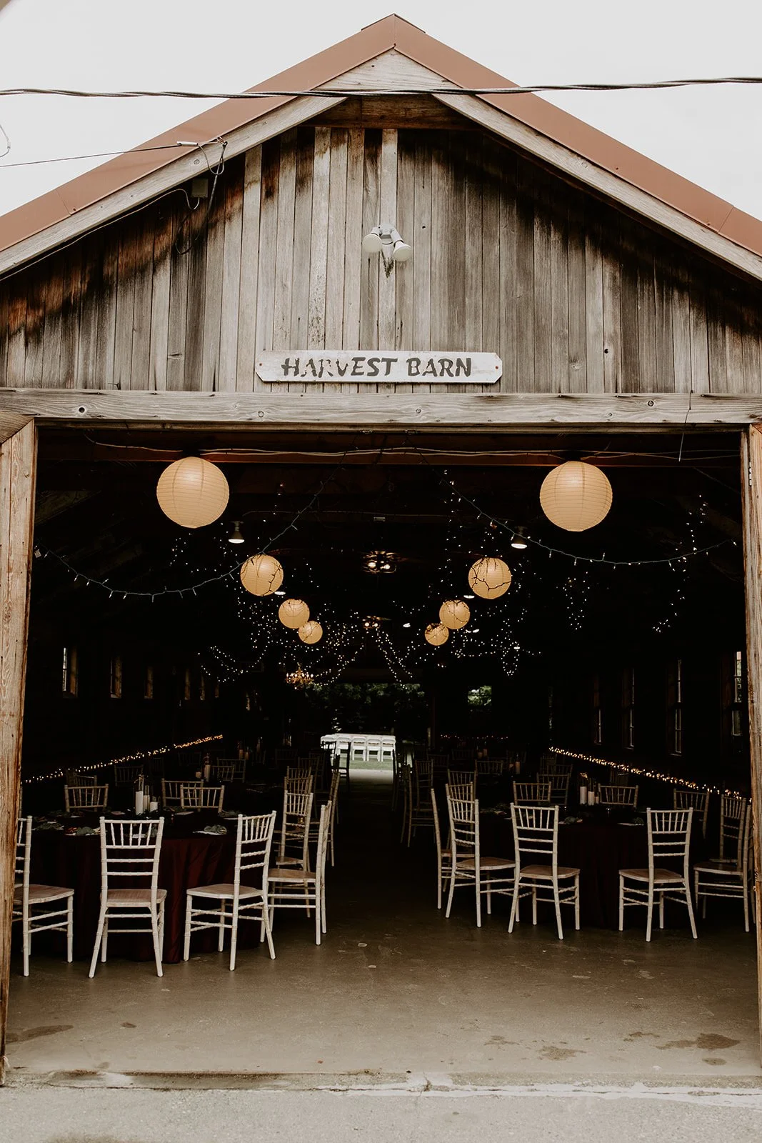 Open wooden barn with string lights and paper lanterns hanging from the ceiling, decorated for an event, with tables and chairs inside. Sign reads "Harvest Barn."