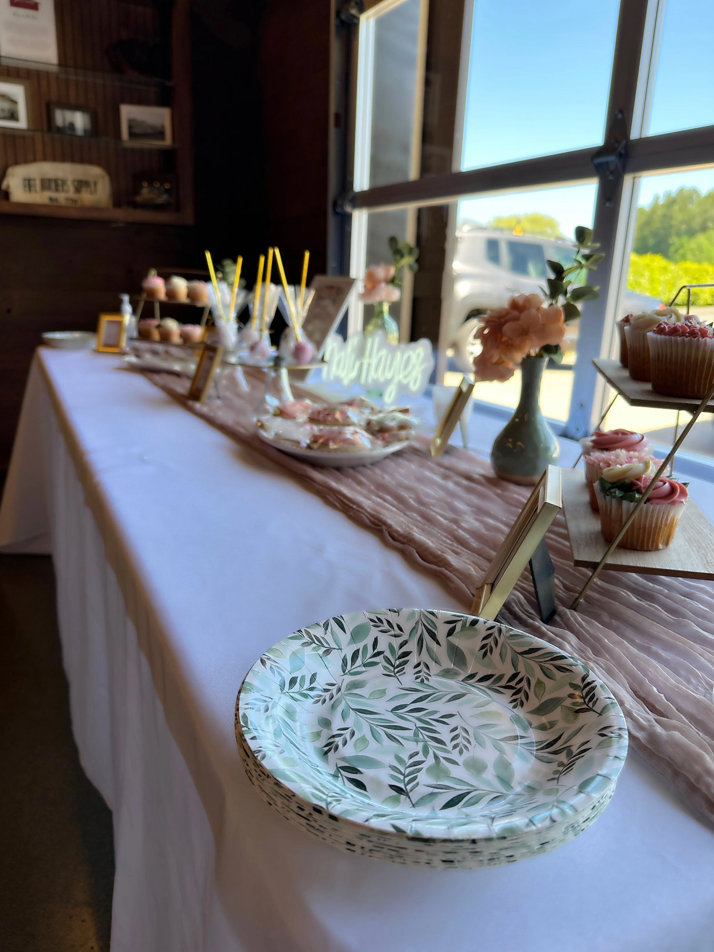 A dessert table decorated with cupcakes, cake pops, and floral arrangements, with a white tablecloth and a pink runner, set near large windows showing a sunny outdoor view.