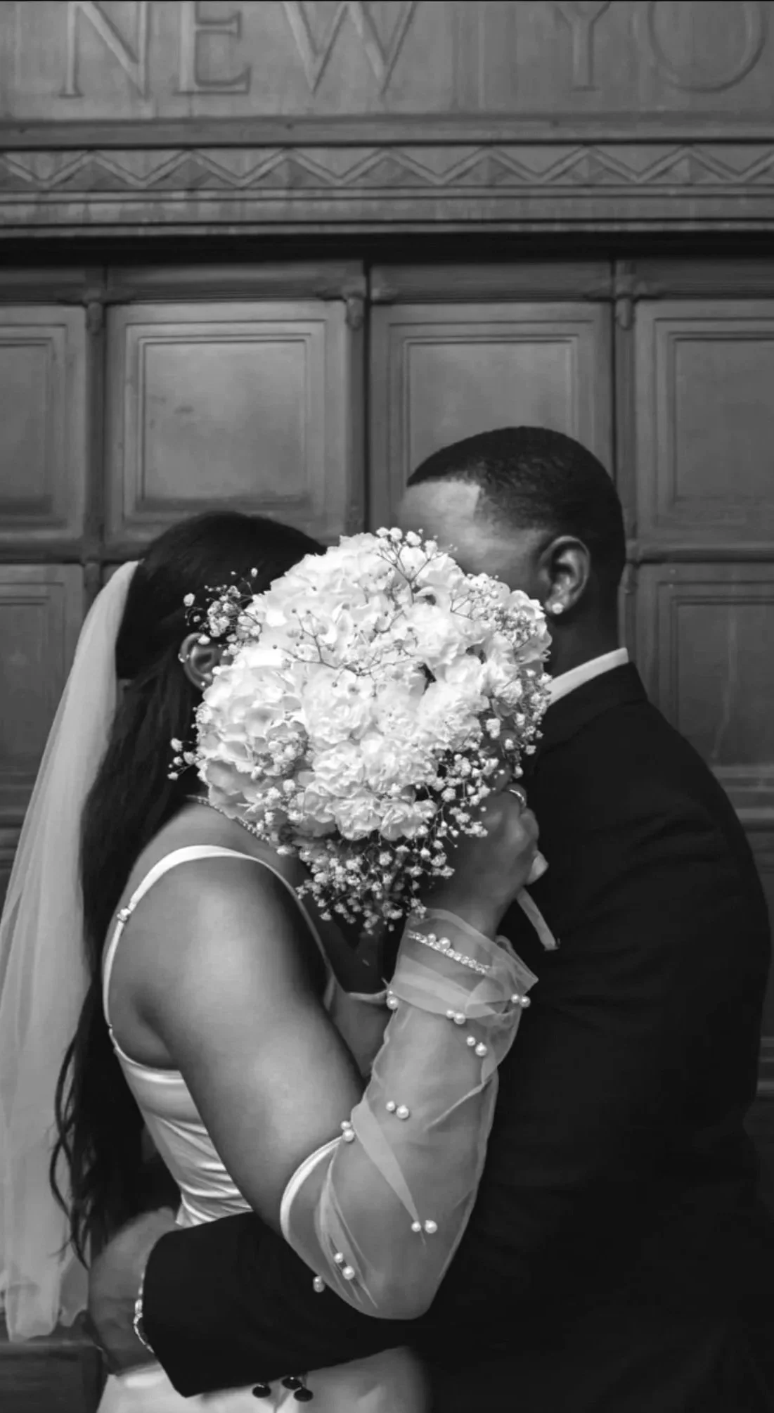 A black and white photo of a bride and groom kissing at their wedding, with the bride holding a large bouquet covering their faces, standing in front of a wooden wall.