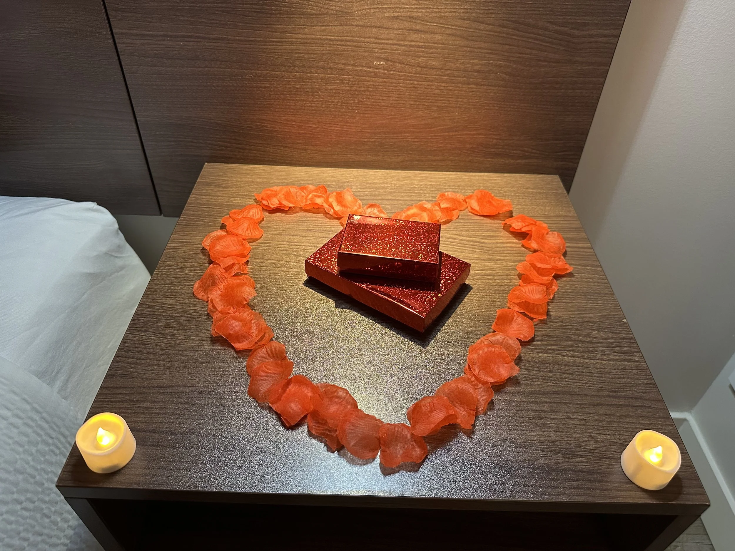 Heart-shaped outline made of orange flower petals on a wooden table with two red gift boxes in the center, illuminated by two small candles at the bottom corners.