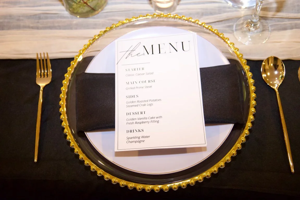 Elegant table setting with a decorative gold beaded charger plate, a white dinner plate, and a menu card on top. Gold fork and spoon placed on a black tablecloth, with a black cloth napkin beneath the menu. Glassware is visible in the background.