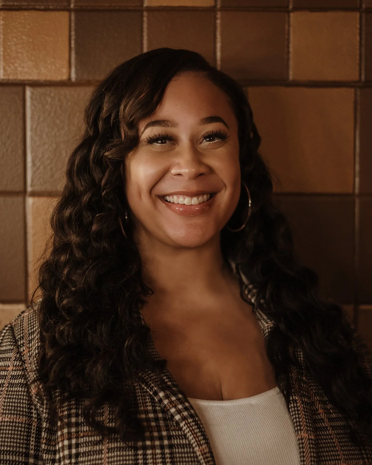 A woman smiling with long curly hair, wearing hoop earrings, a plaid blazer, and a white top, standing against a brick wall.