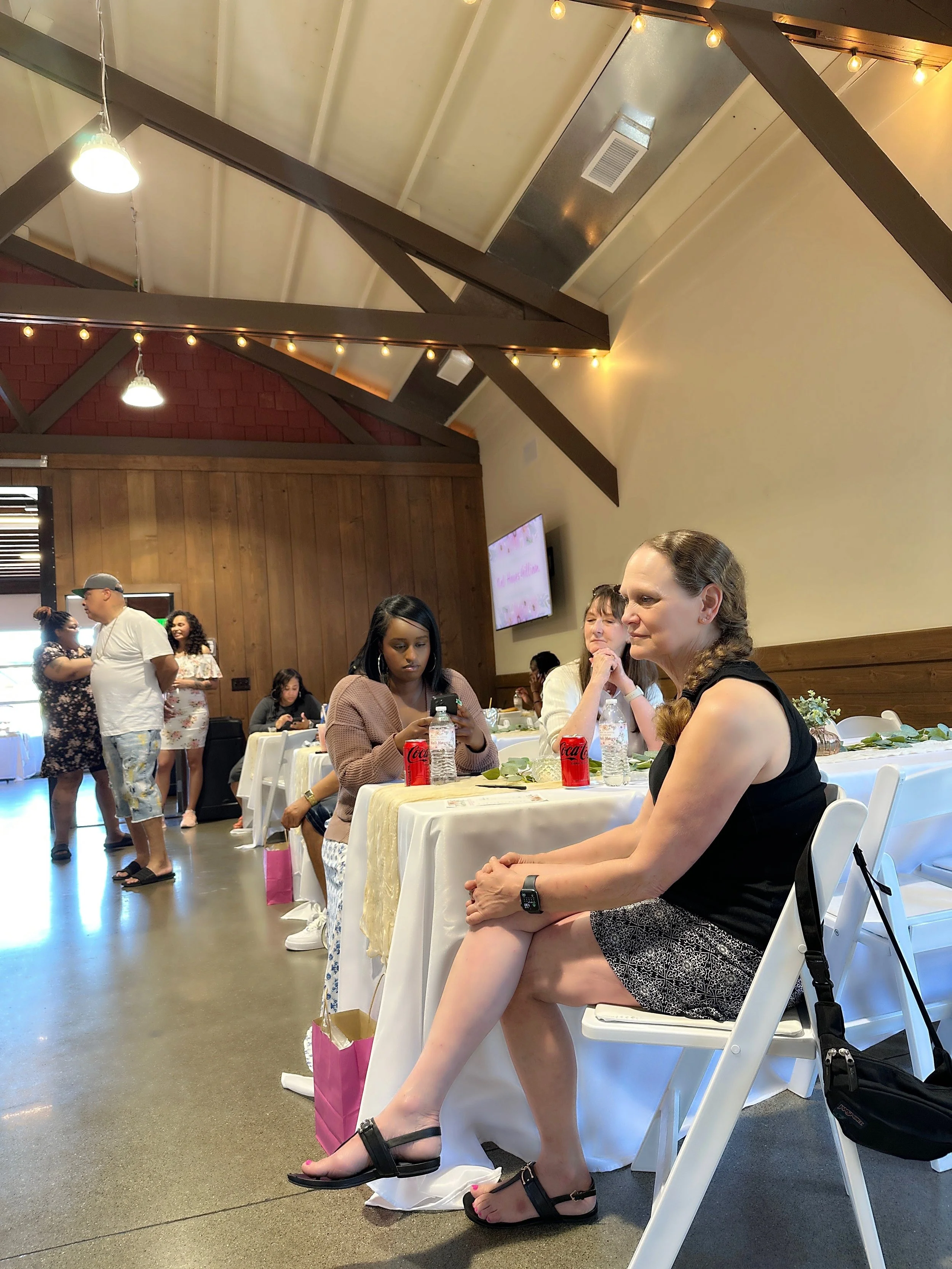 People sitting at tables during a social event or celebration in a room with wooden walls and a vaulted ceiling with string lights.