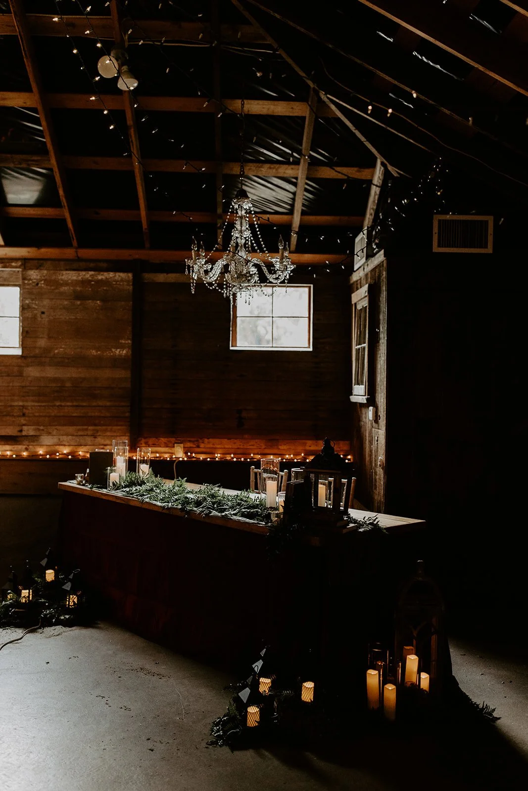 Interior of a rustic barn decorated for a cozy event with string lights, candles, and a crystal chandelier.
