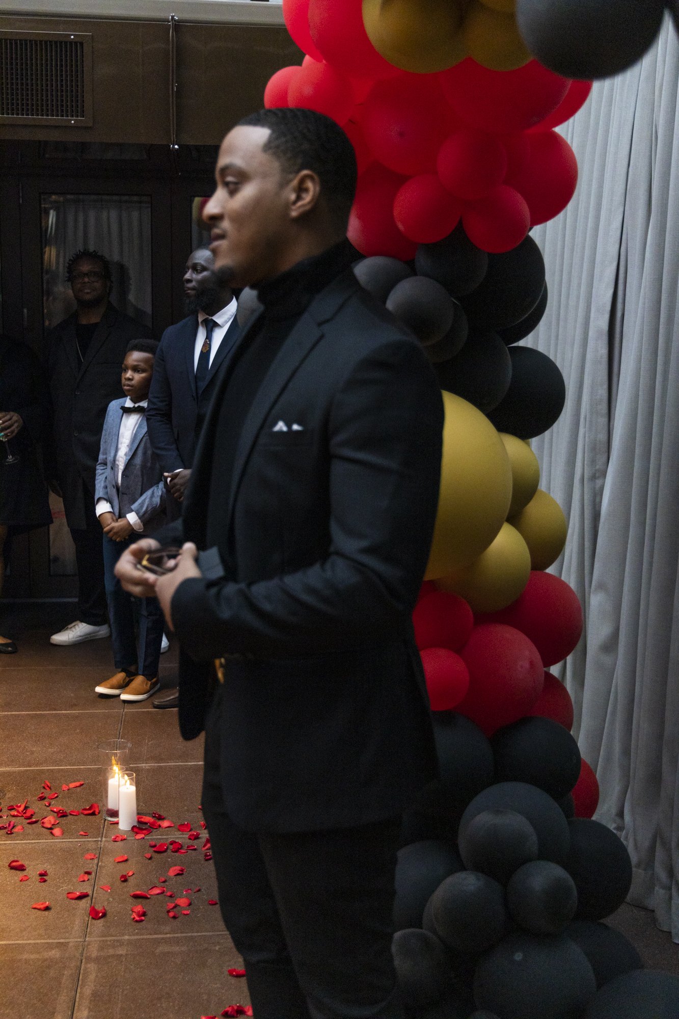 Man in a black suit standing near a colorful balloon arch at an indoor event.
