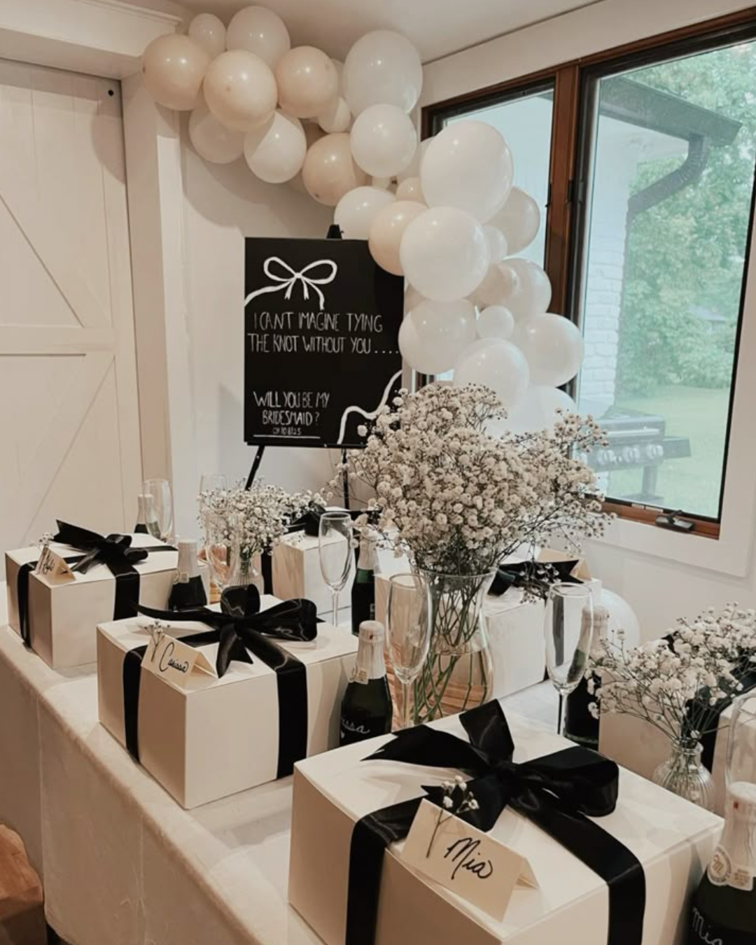 Table with white gift boxes decorated with black ribbons, champagne bottles, champagne flutes, white floral arrangements, and a black sign with white text, surrounded by white balloons, set near a window with greenery outside.