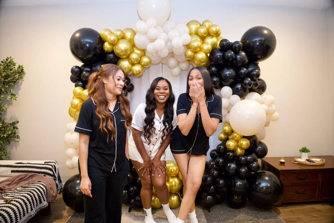 Three young women in pajamas standing in front of a balloon arch with black, white, and gold balloons, appearing happy and laughing.