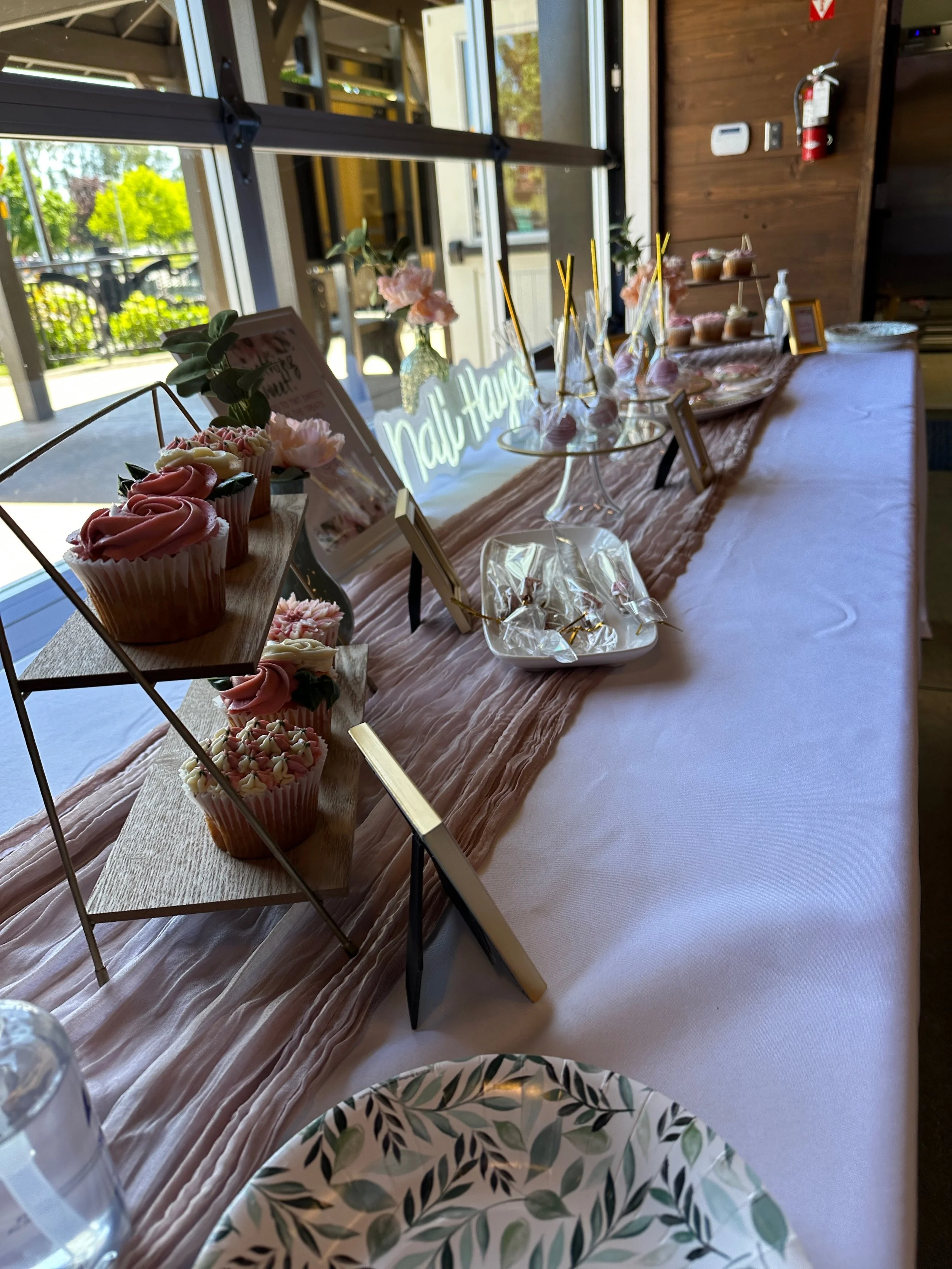 A dessert table with pink cupcakes, flower decorations, and a neon sign reading 'Nail Happy,' set near a large window with outdoor greenery visible.
