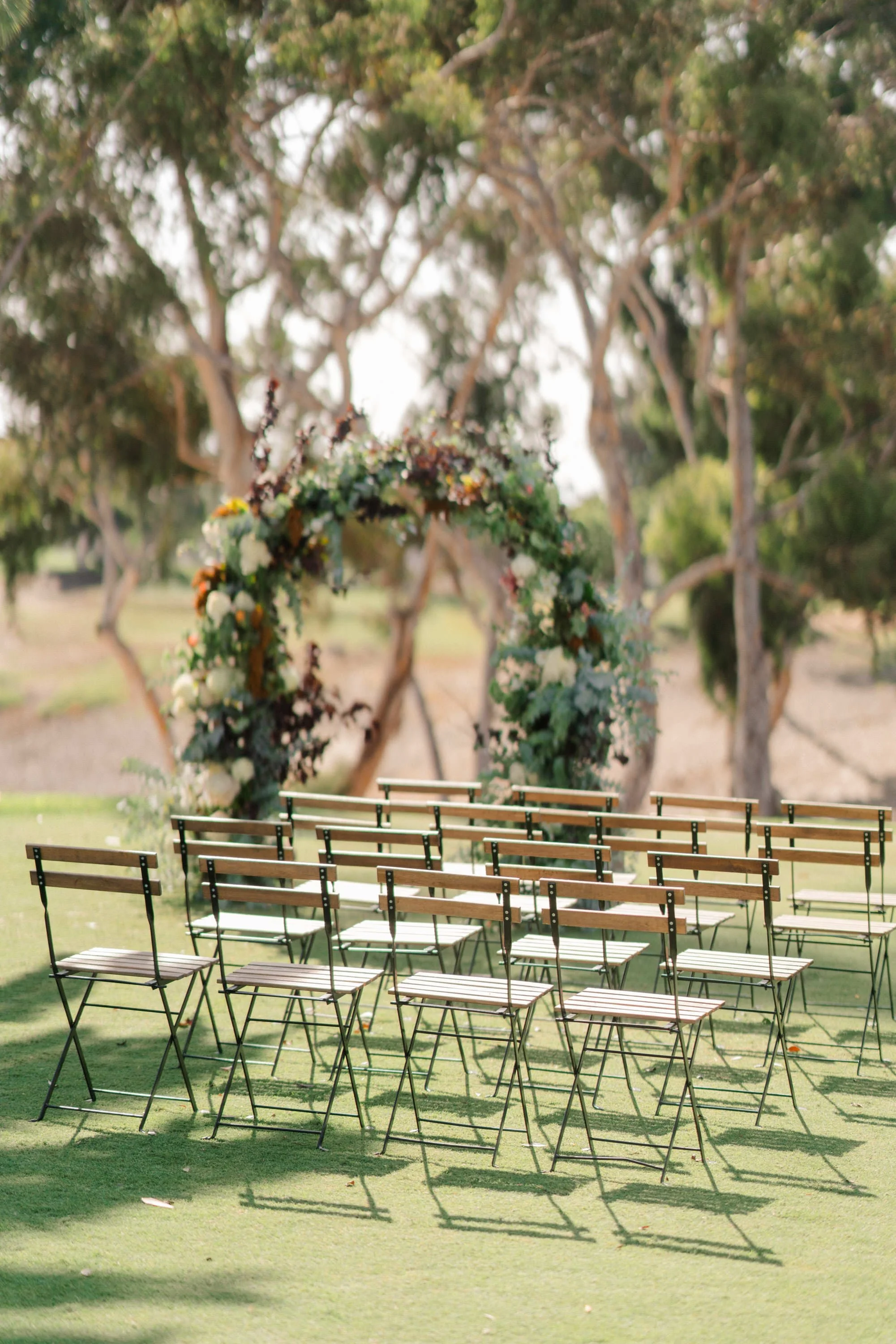Outdoor wedding ceremony setup with multiple wooden chairs arranged on green grass facing a floral arch in a park.