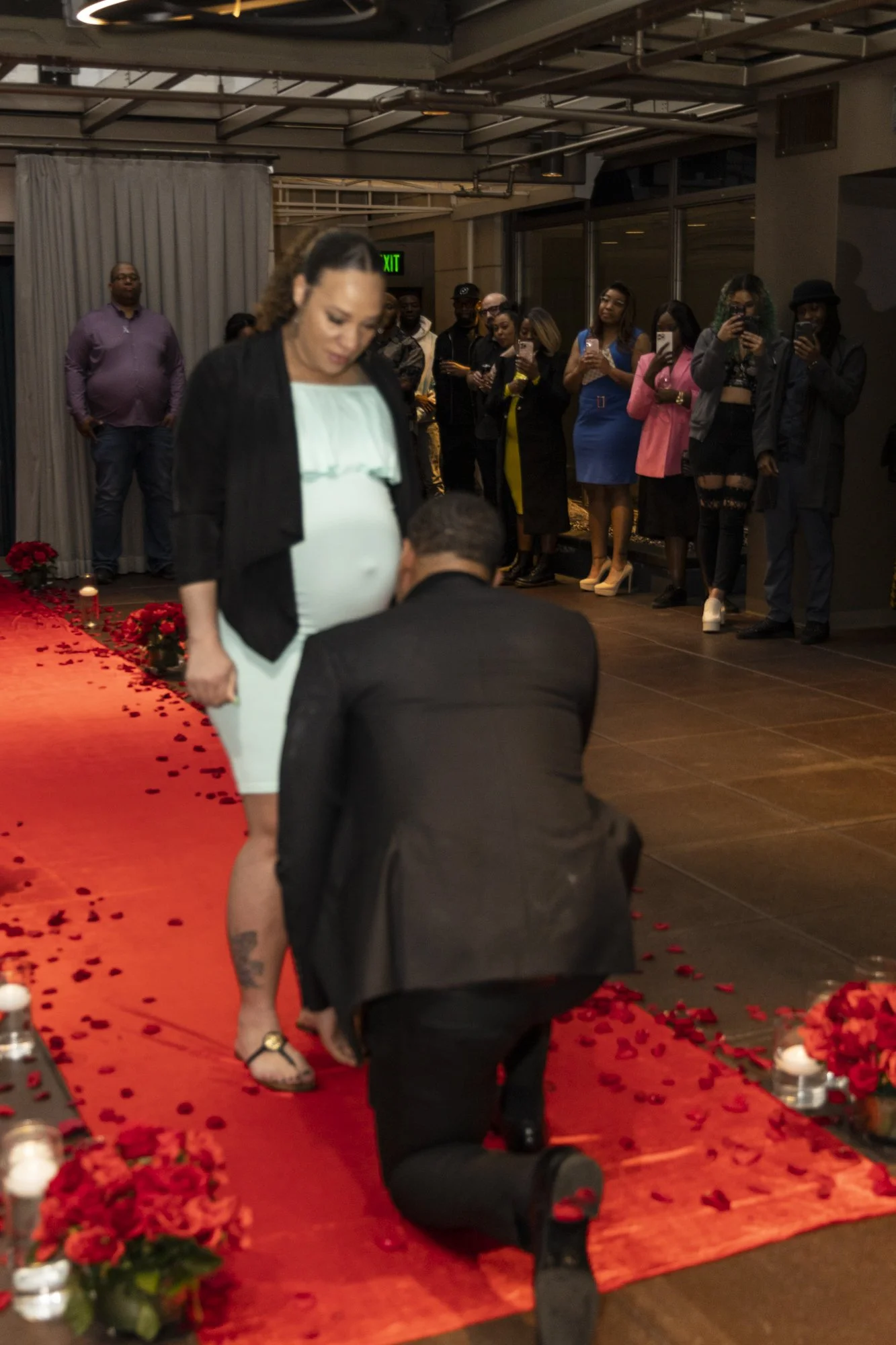 A man proposes marriage to a pregnant woman during a romantic setting with red carpet and flower decorations, while friends and family watch and take photos.